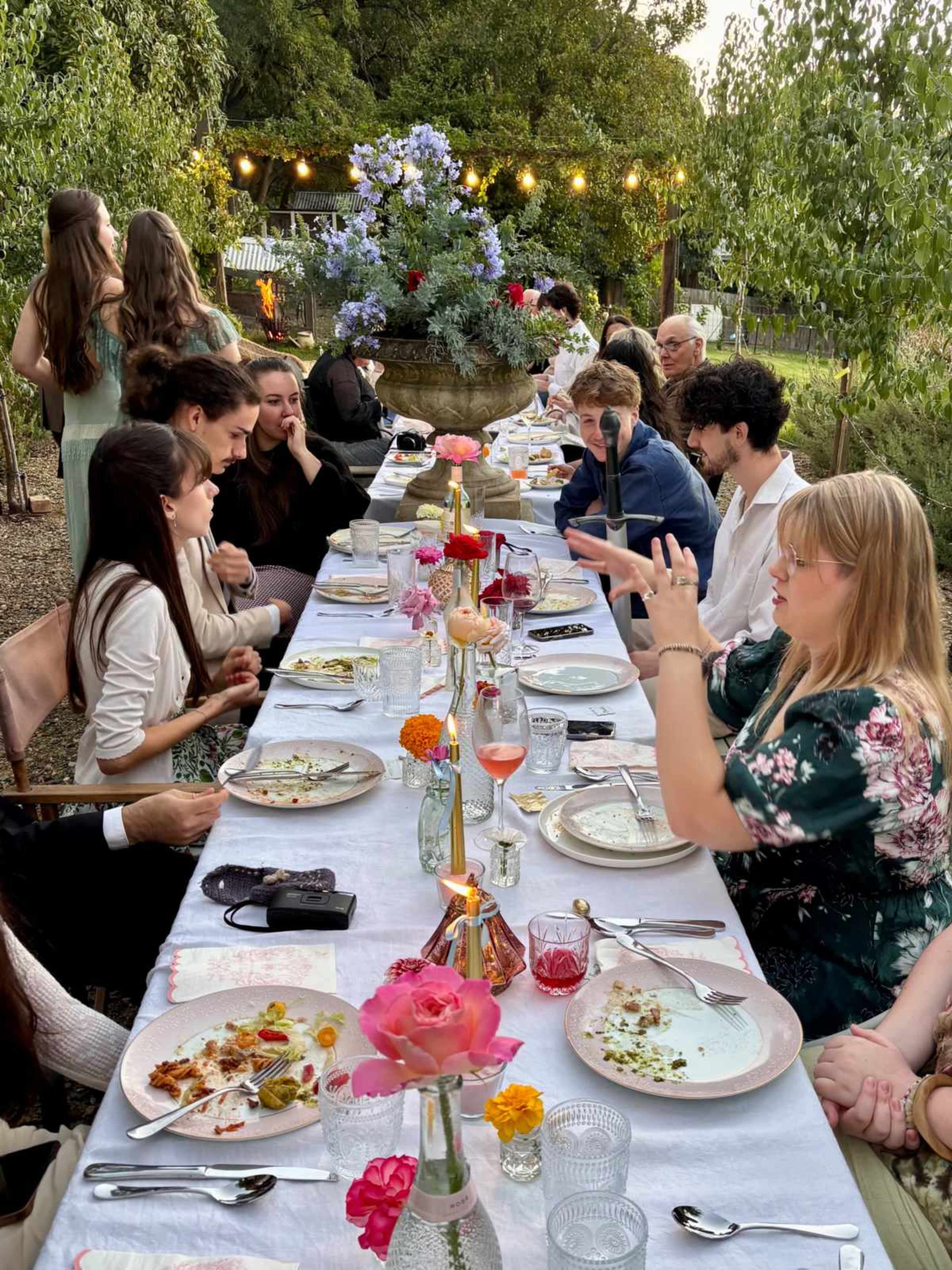 A long table is set outdoors for a gathering, with guests seated and engaged in conversation, surrounded by greenery and decorative lights.