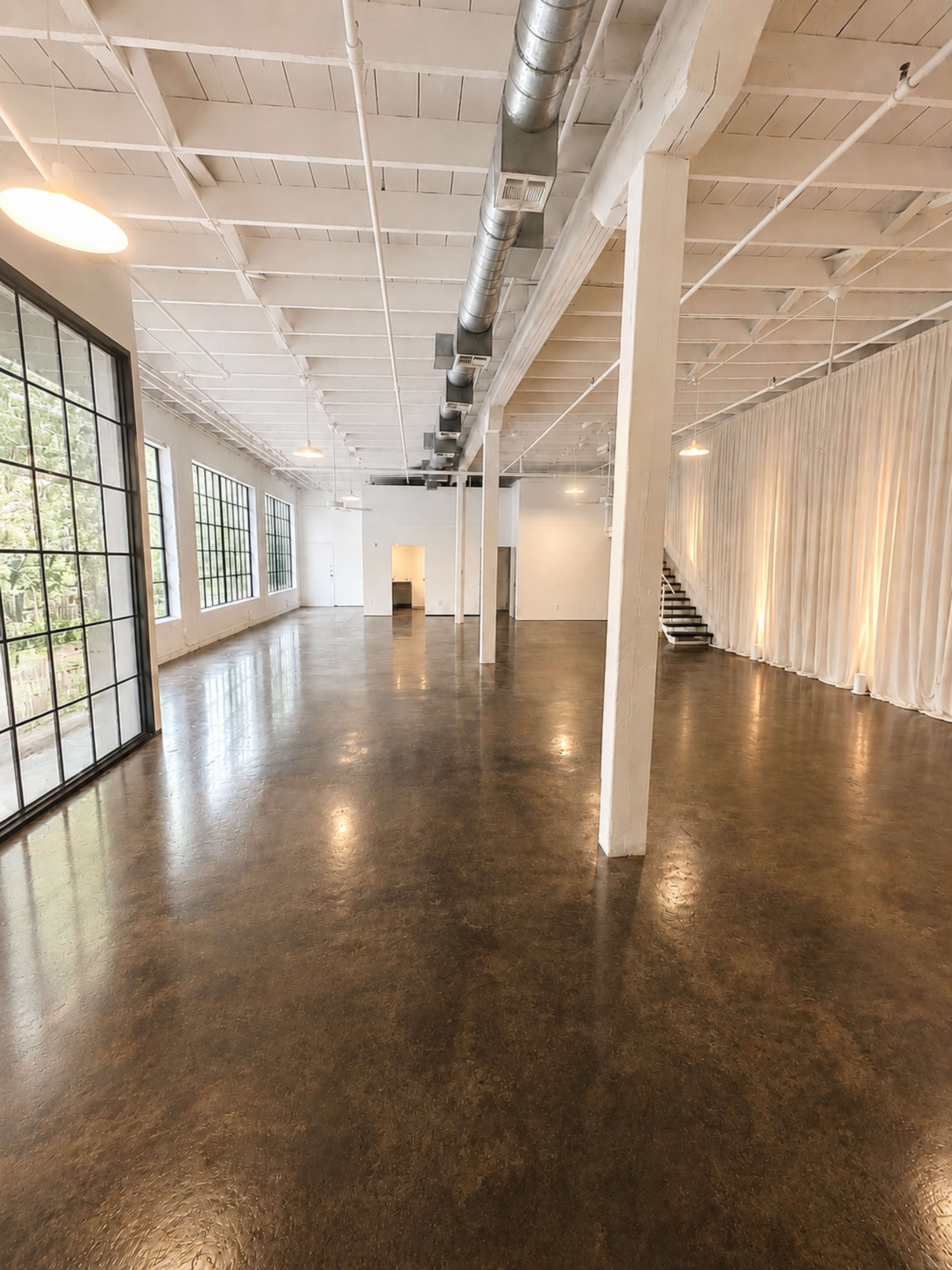 The image shows a spacious, empty room with high ceilings, large windows, polished concrete floors, and soft lighting along one wall.