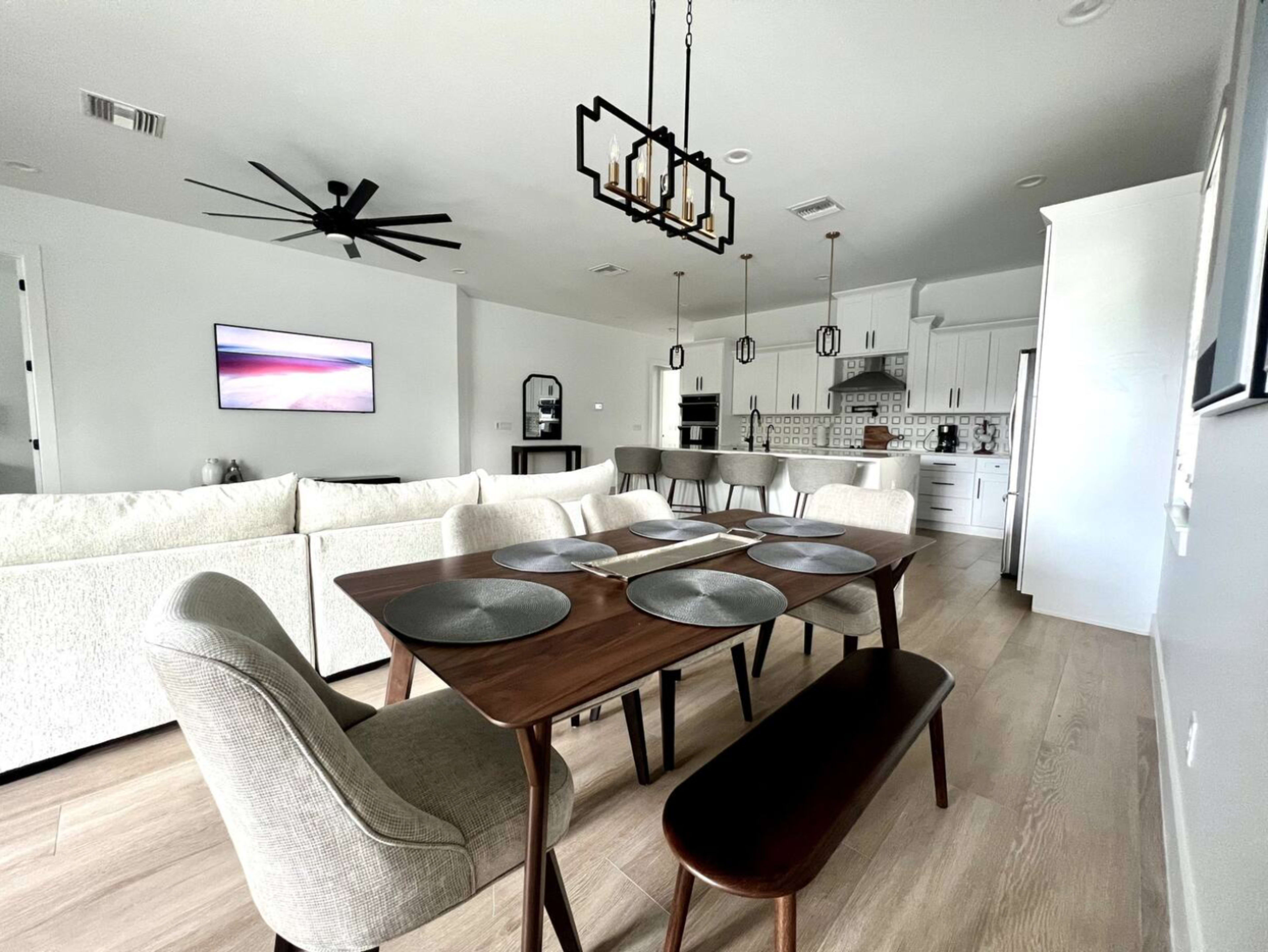 The image shows a modern dining area with a wooden table set for six, surrounded by upholstered chairs and a bench, with an adjoining kitchen visible in the background.