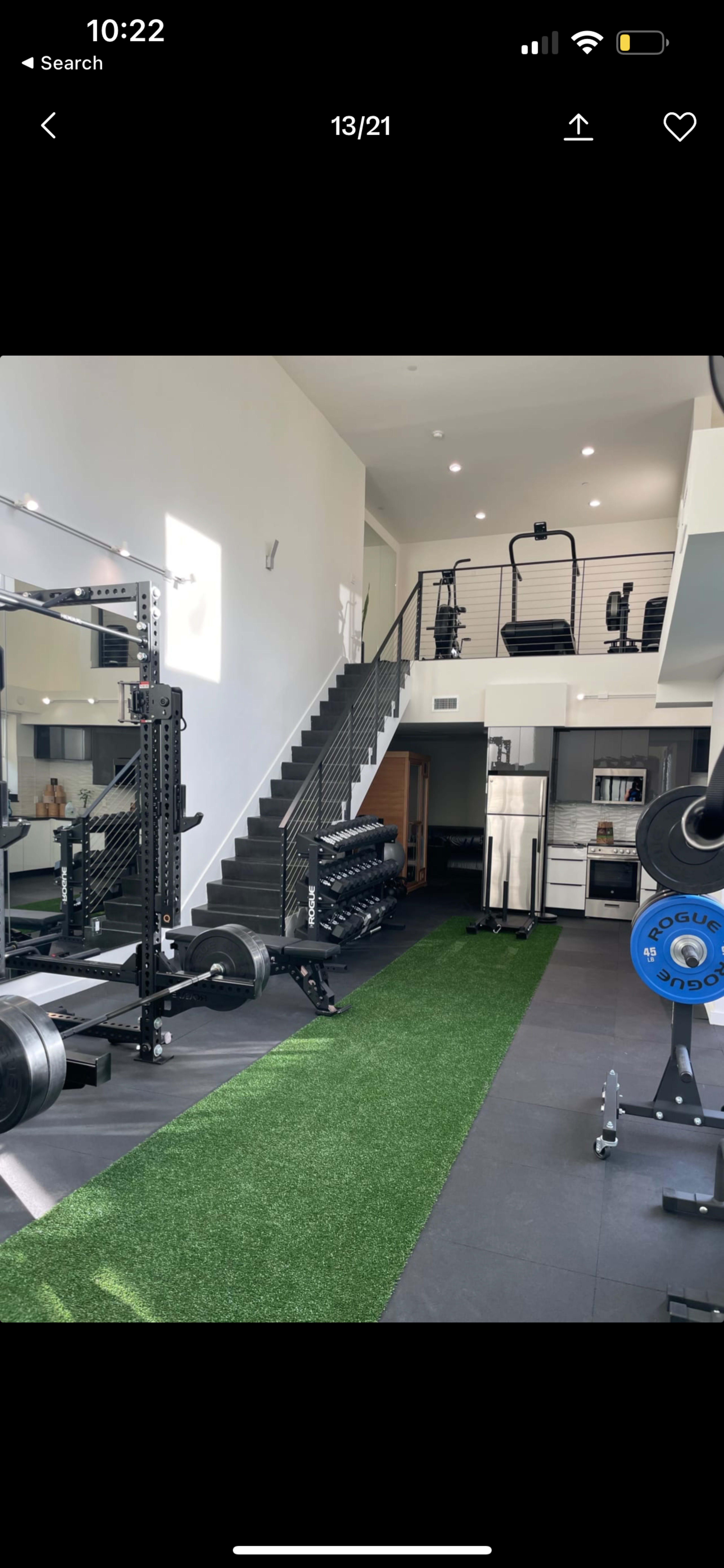 A modern gym interior featuring weightlifting equipment, a green turf aisle, and a staircase leading to an upper level.
