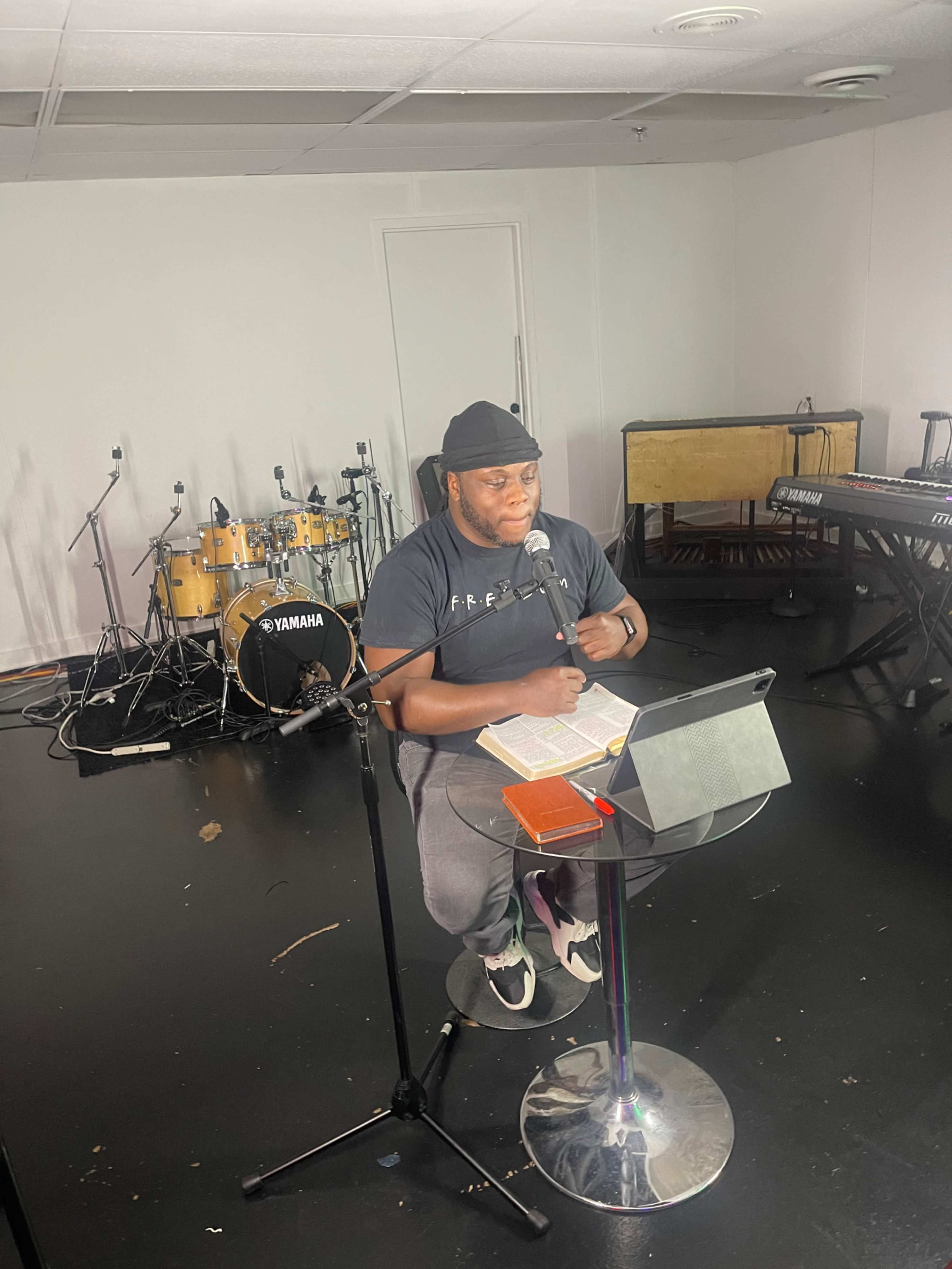A man sits at a microphone in a music studio, reading from a book, with drums and keyboards visible in the background.