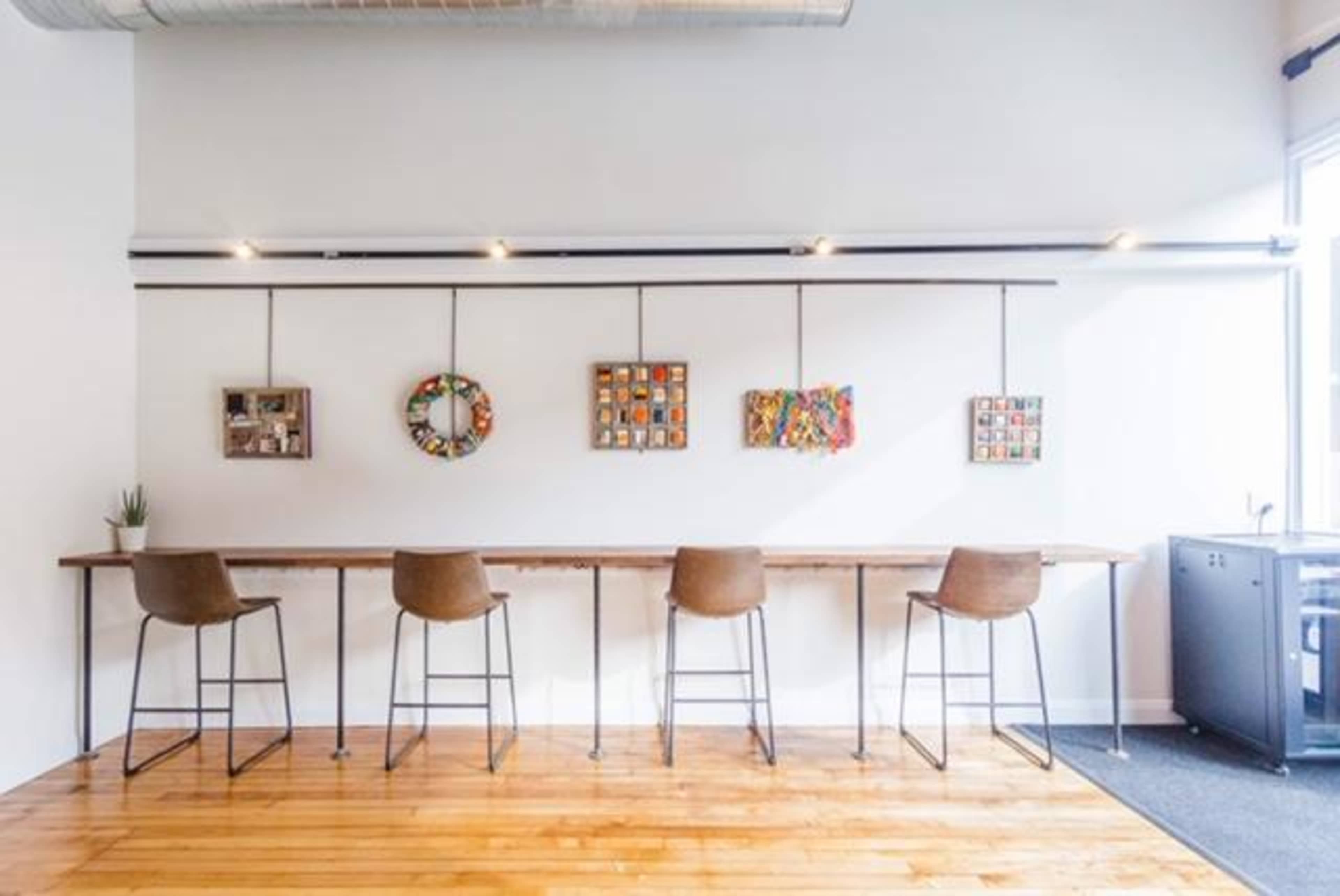A modern workspace featuring a long wooden table with four stools against a wall adorned with five framed art pieces.