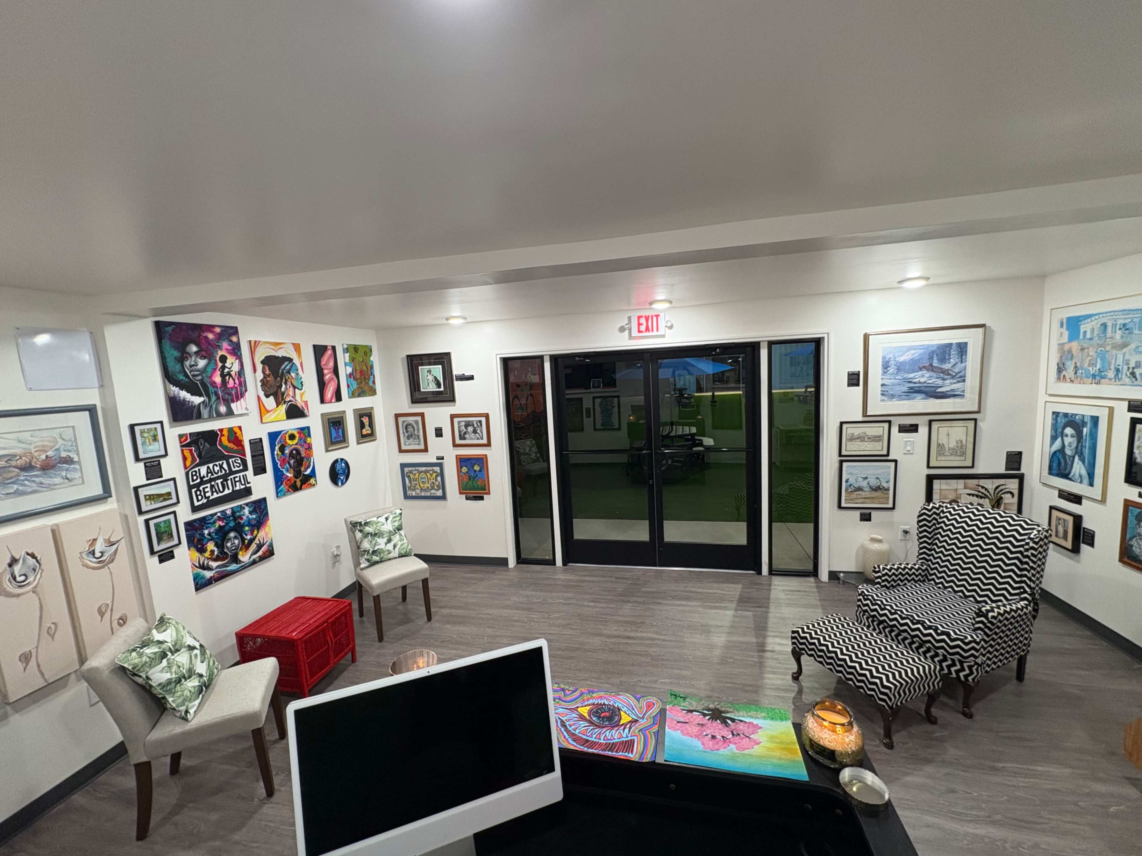 The image shows an interior room decorated with various pieces of artwork on the walls, a patterned armchair, and a visible exit door.