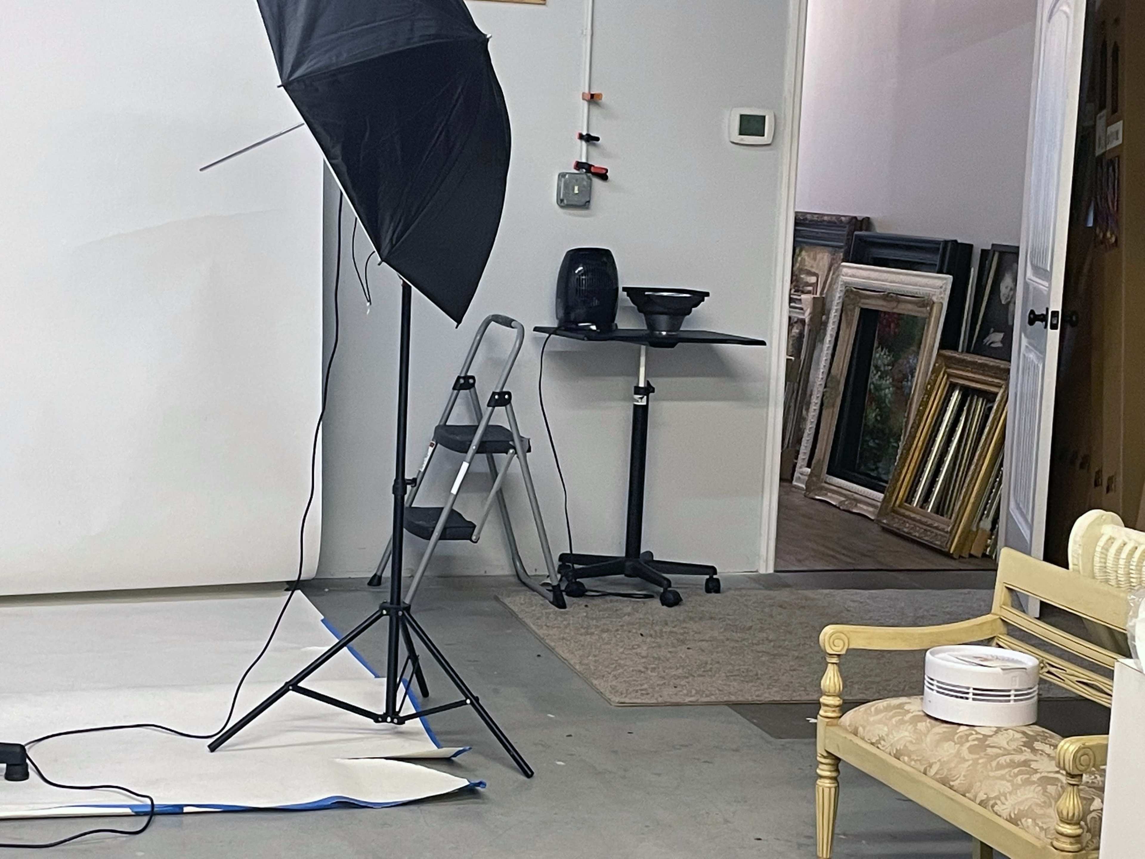 The image shows a photography studio setup with a softbox light, a small folding ladder, a rolling cart, and an open door revealing a room with framed artwork.