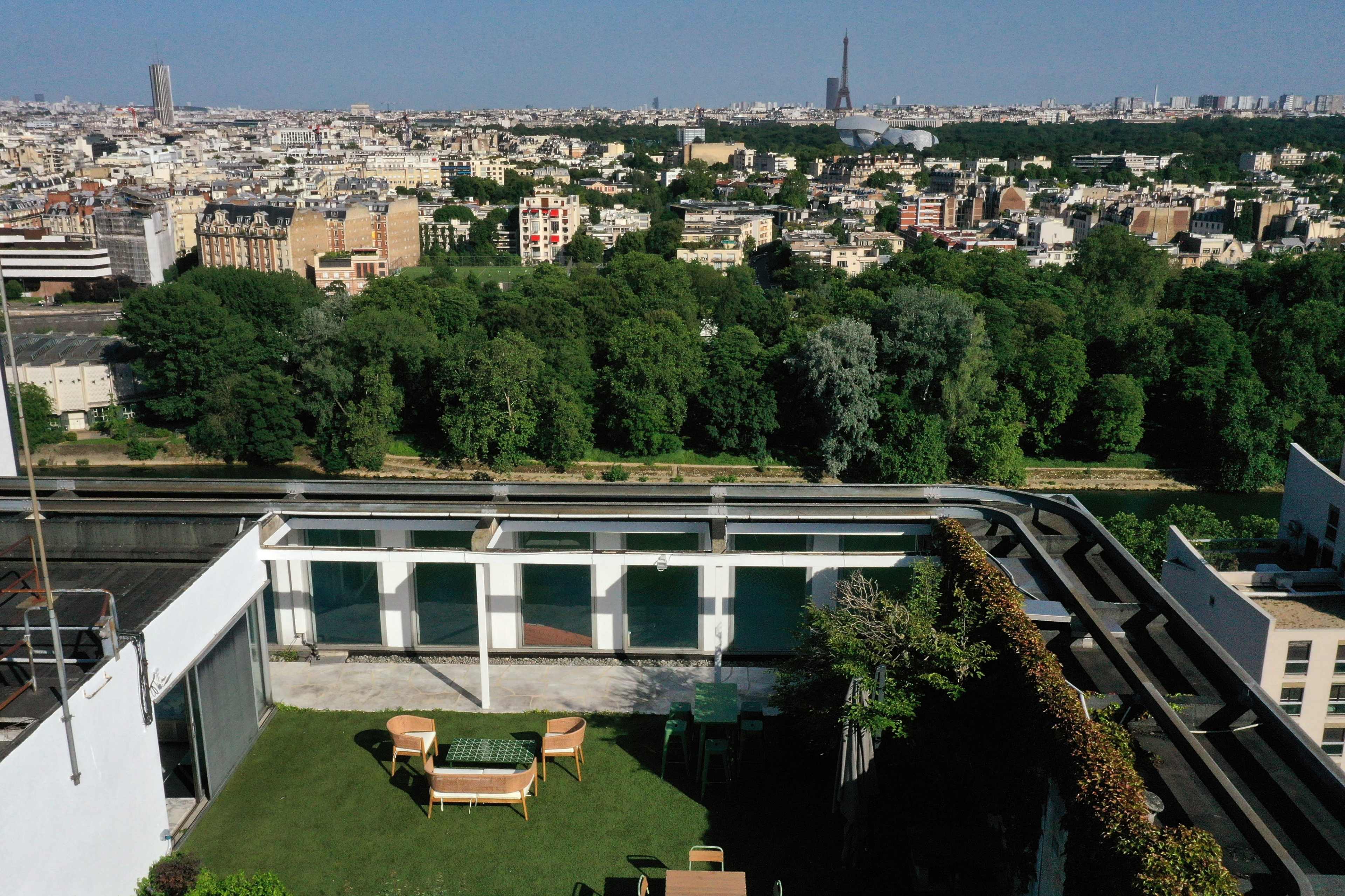 The image shows a rooftop terrace with green grass and wooden furniture overlooking a cityscape with buildings and a park in the background.