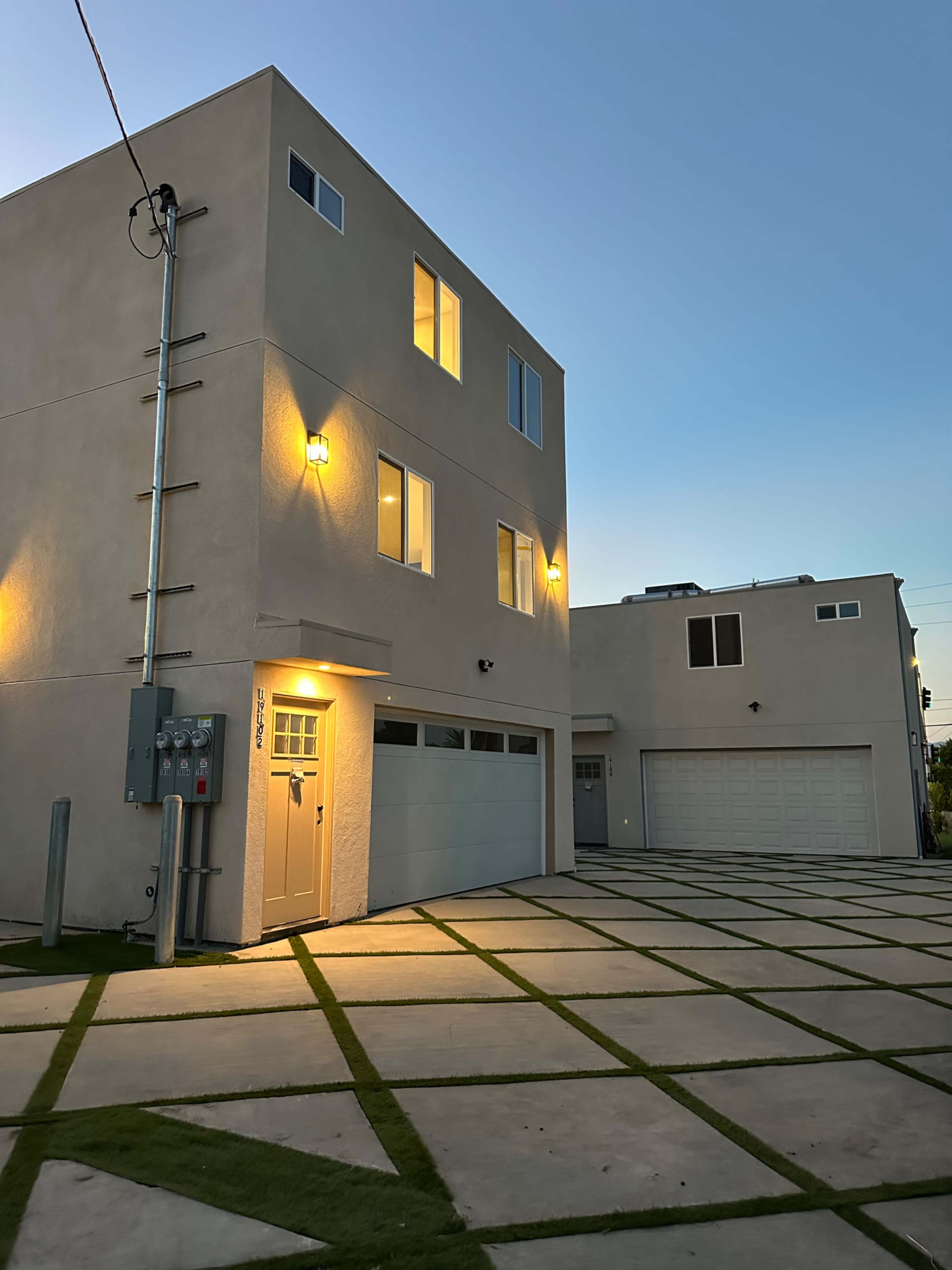 The image shows a modern two-story building with illuminated windows and a garage, situated next to another similar structure on a paved area with grass in between.