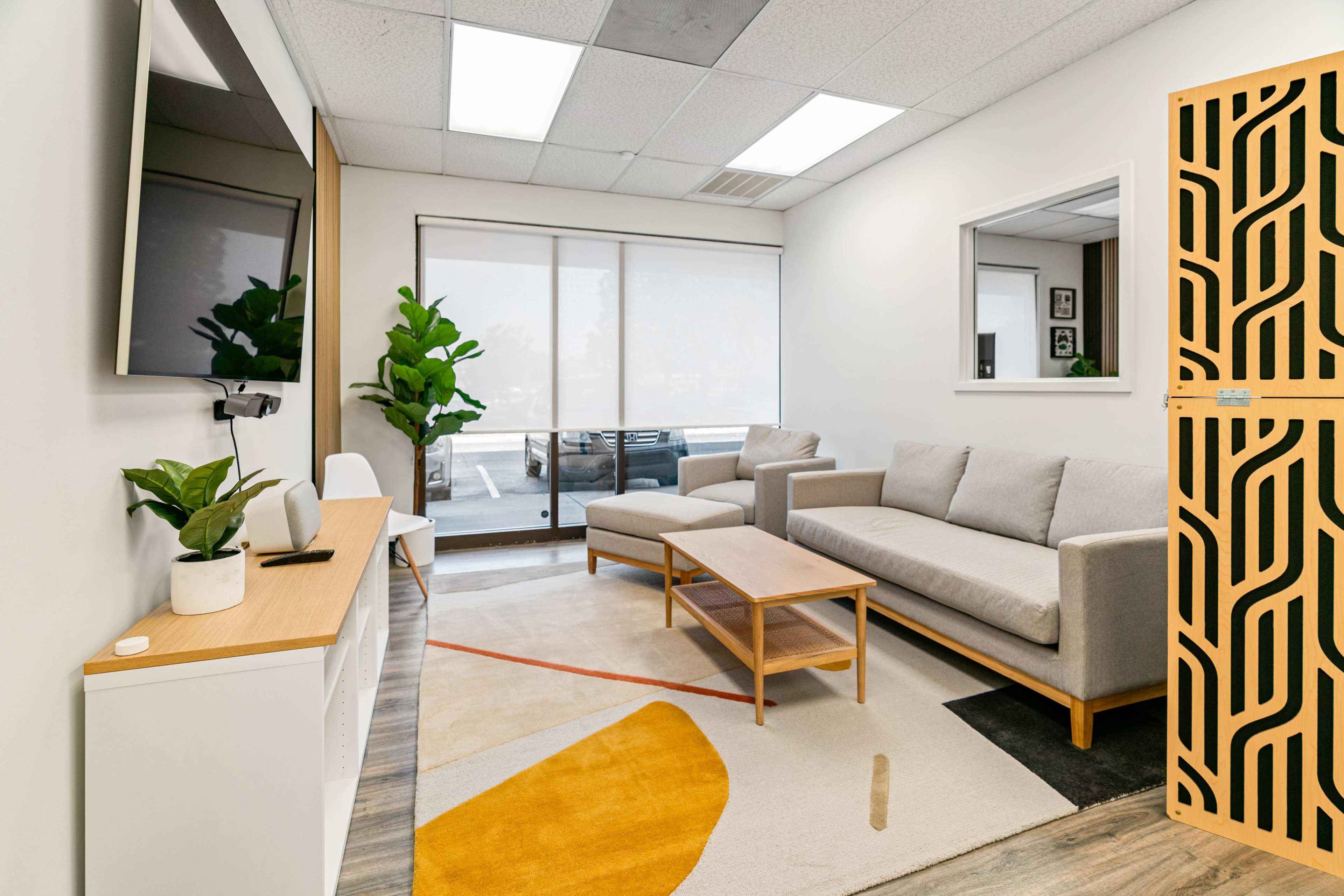 A modern waiting area featuring a gray sofa, a wooden coffee table, and a large television against a white wall, with a window displaying outdoor seating and indoor plants.