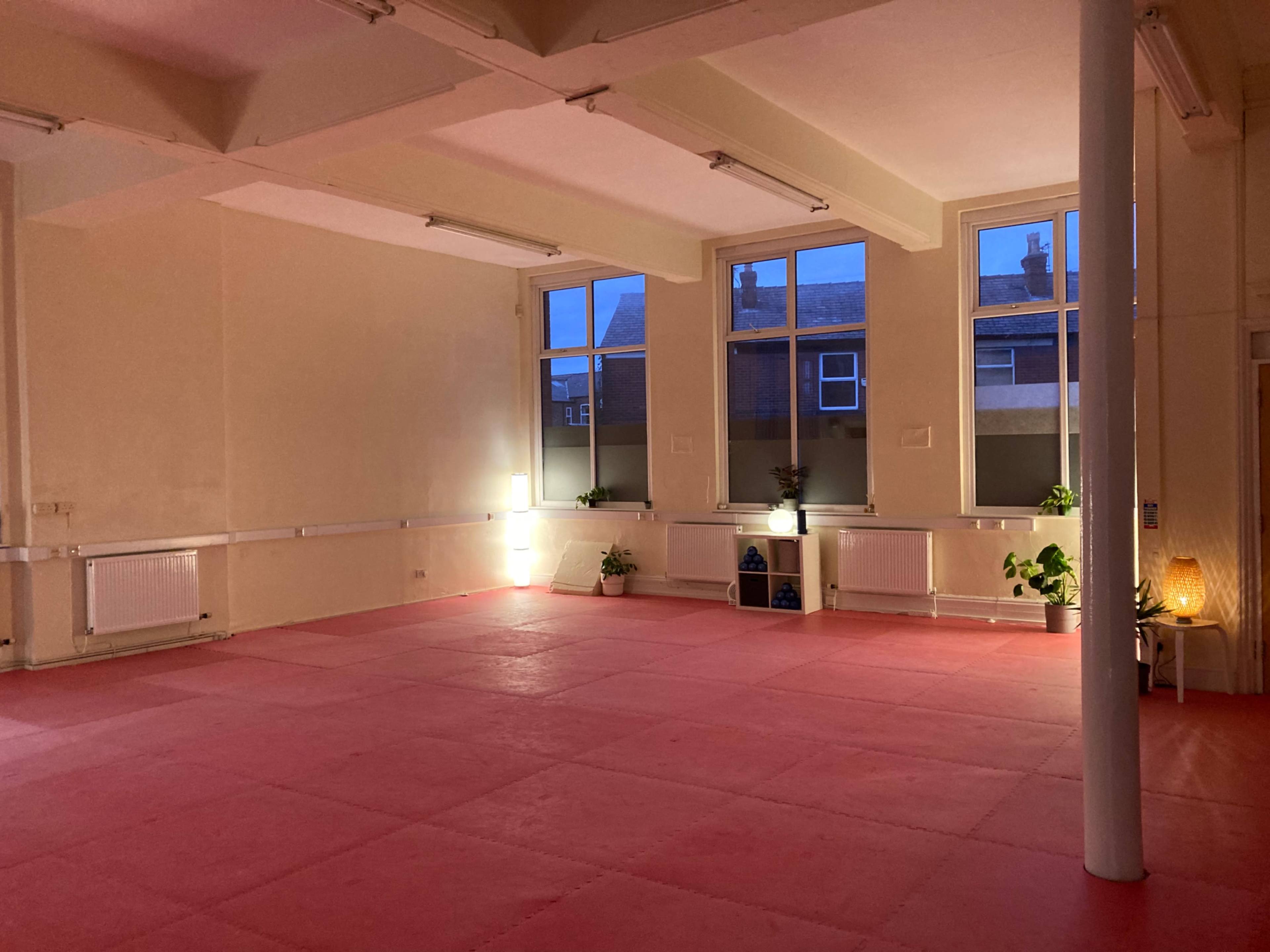 A spacious, empty room with pink flooring, large windows, and minimal lighting, featuring several potted plants.