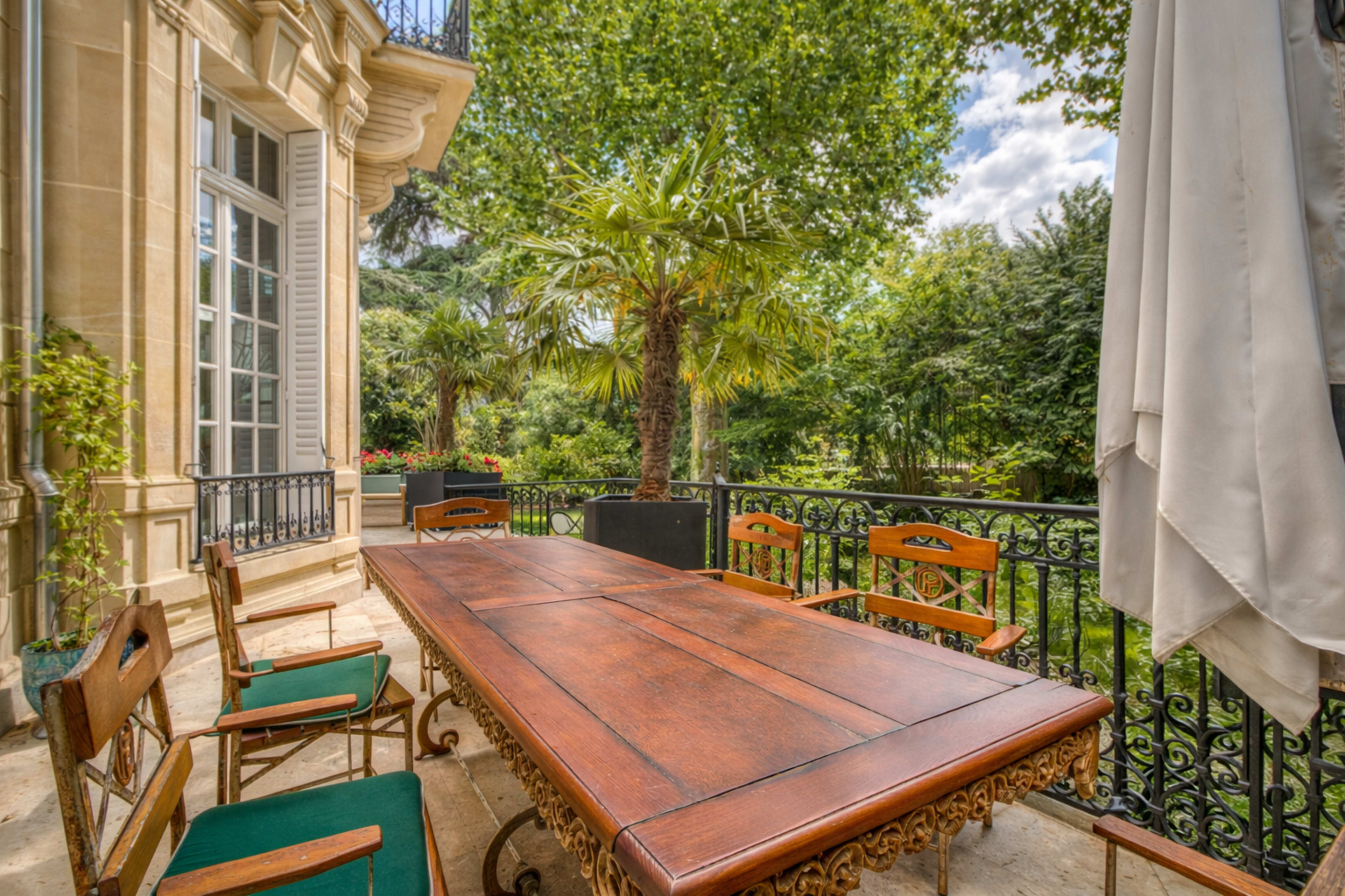 A wooden dining table surrounded by chairs is positioned on a patio overlooking a lush garden with palm trees and greenery.