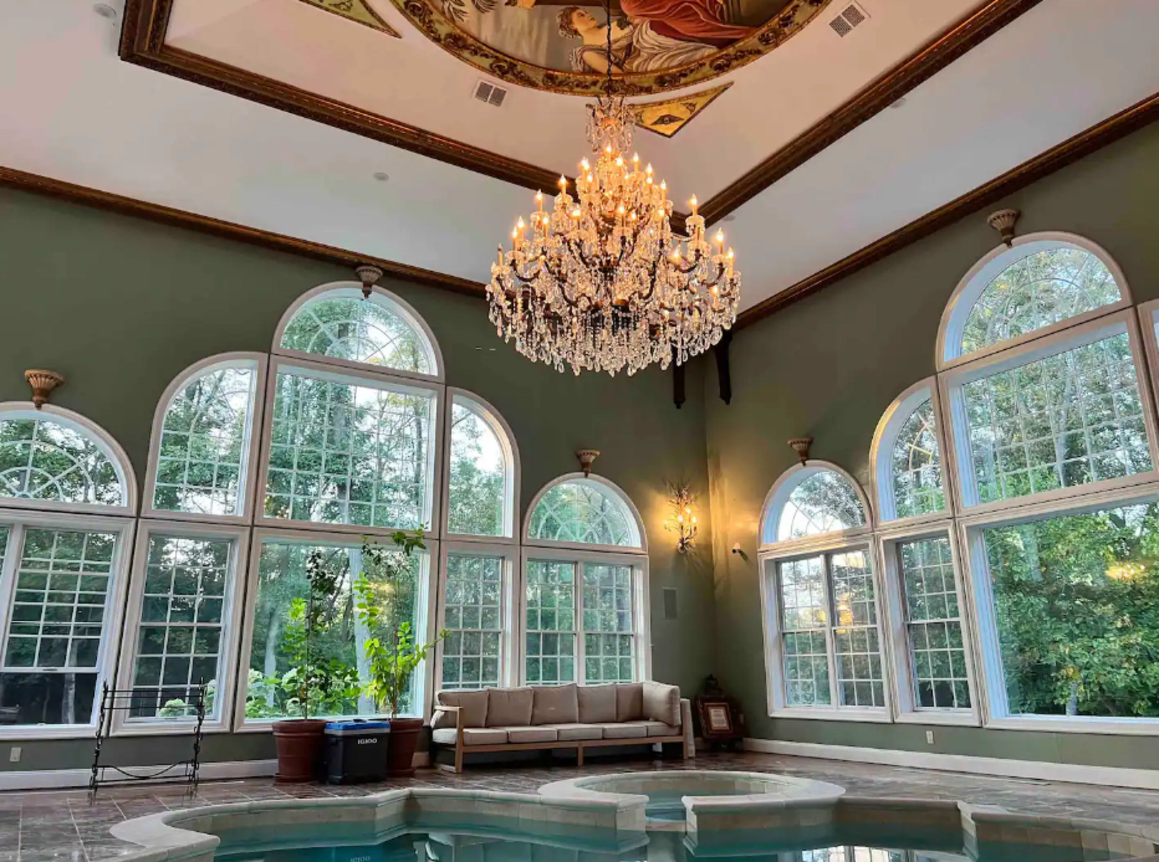 A room with a chandelier, large arched windows, and a small indoor pool surrounded by green walls and a seating area.