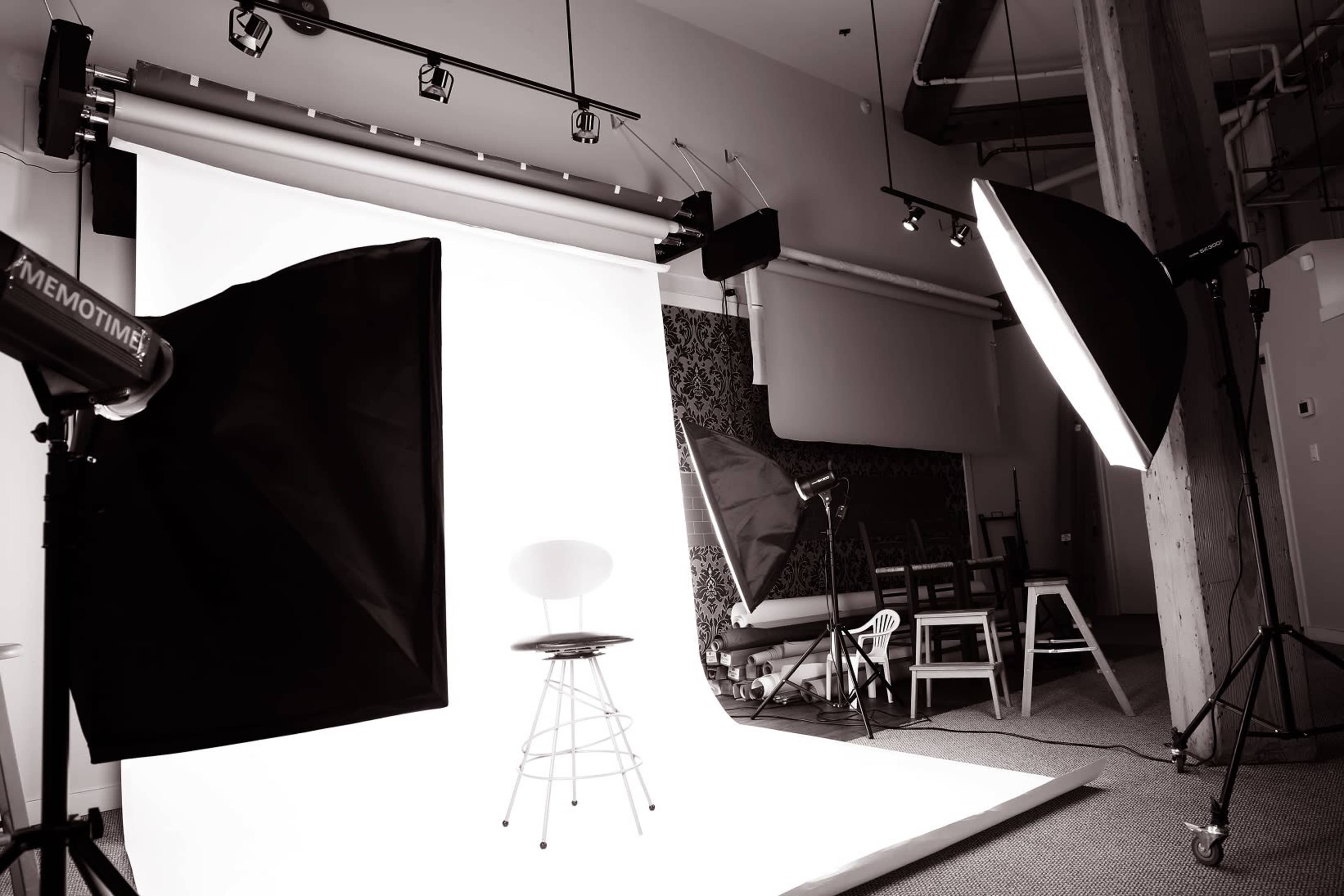 The image shows a photography studio setup with a white backdrop, various lighting equipment, and an empty stool in the center.