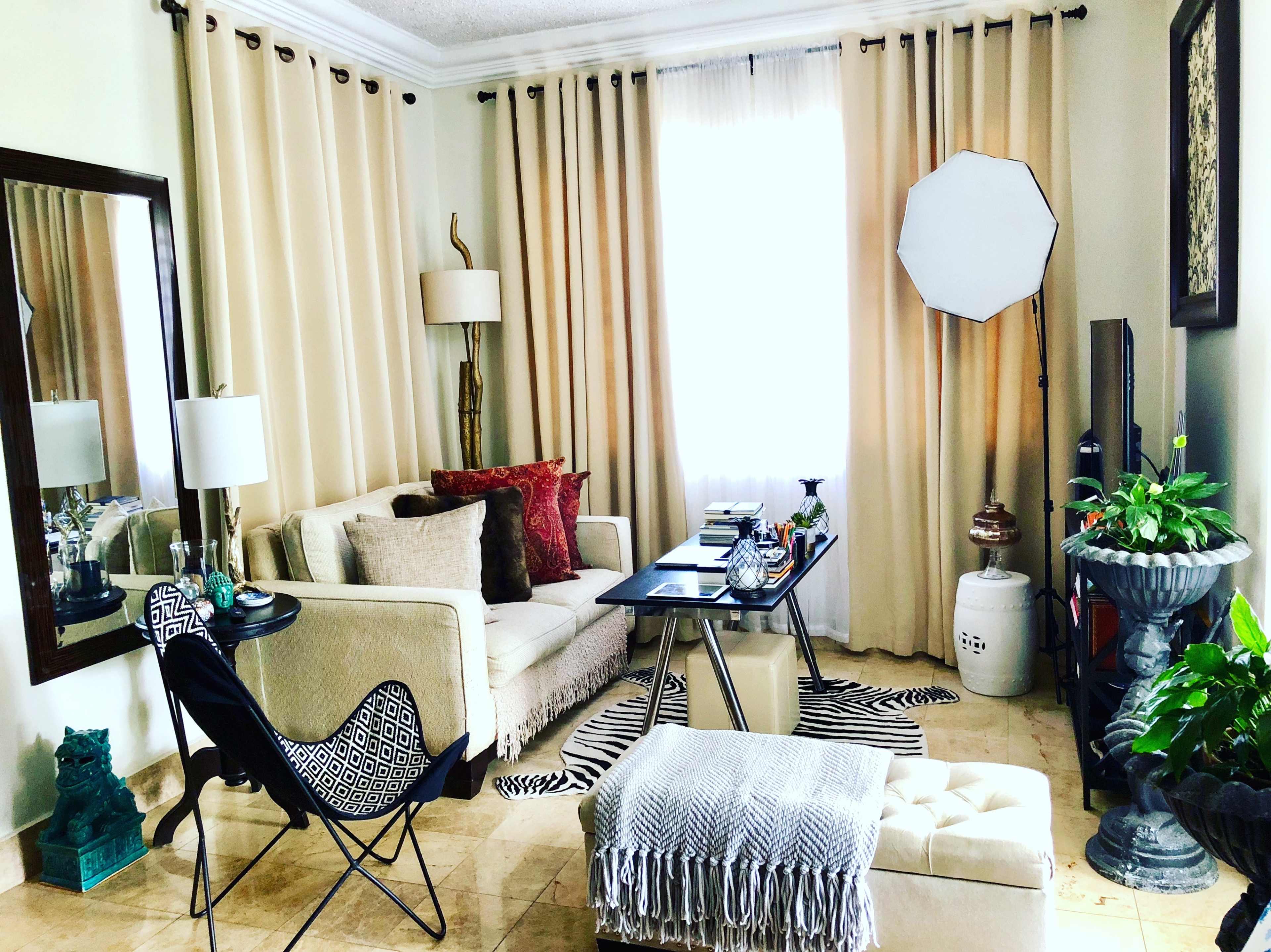 The image shows a living room with a beige sofa, a glass coffee table, a floor lamp, and a large window covered by cream-colored curtains.