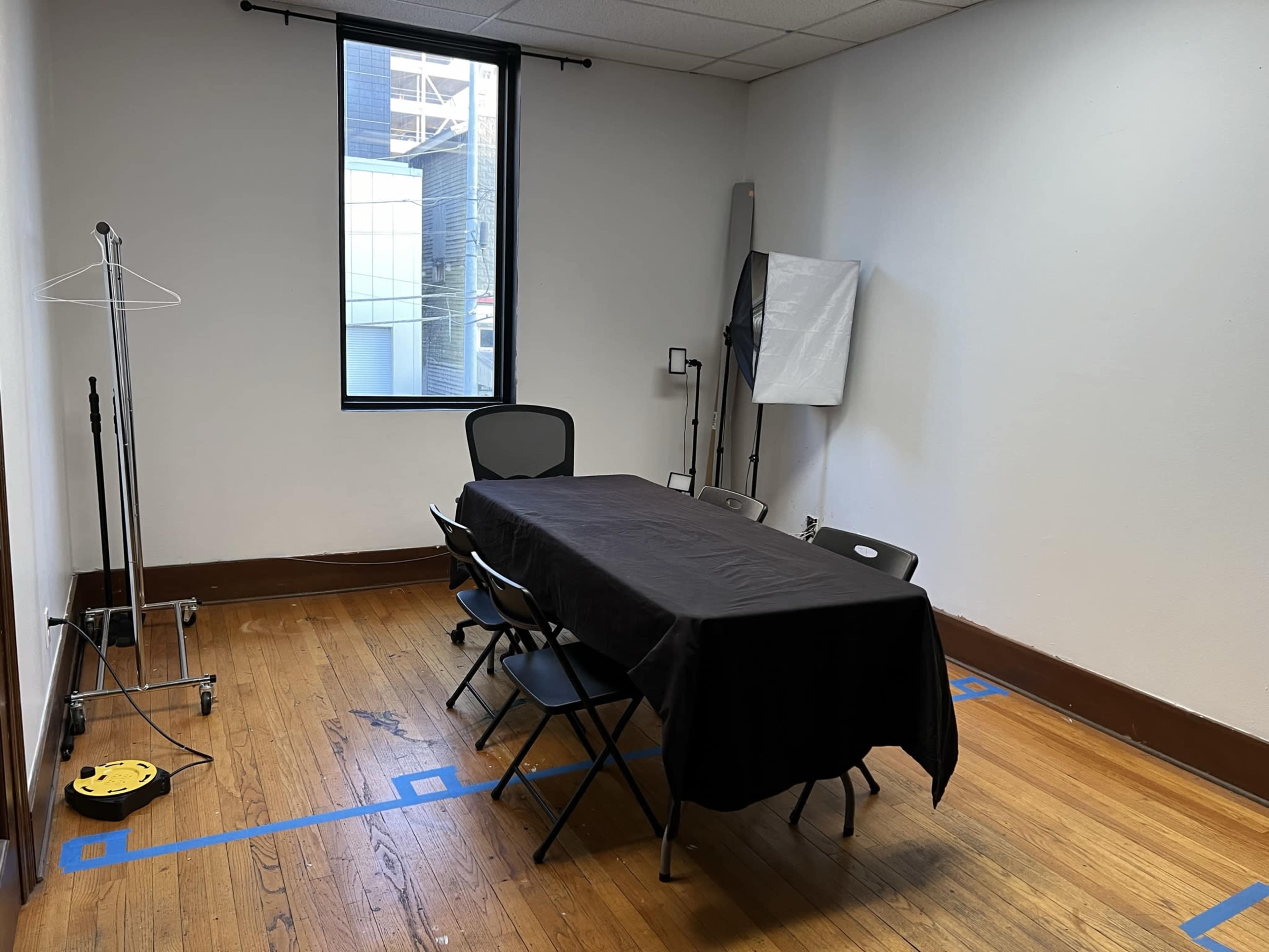 A small, empty room features a table covered with a black cloth and surrounded by several folding chairs, with a window allowing natural light and a clothes rack in one corner.