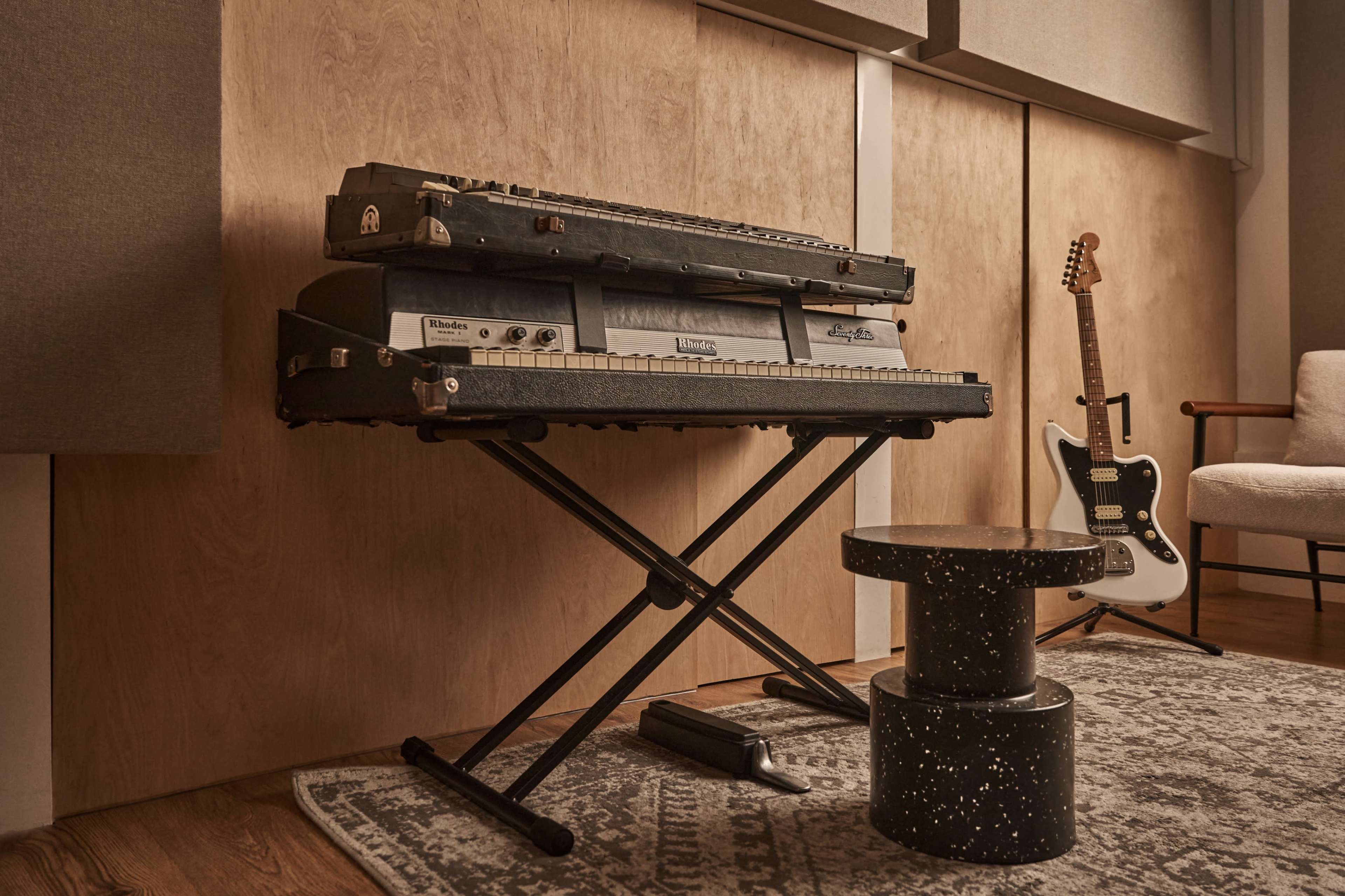 A vintage keyboard sits on a stand next to a modern electric guitar and a small table in a sound studio.