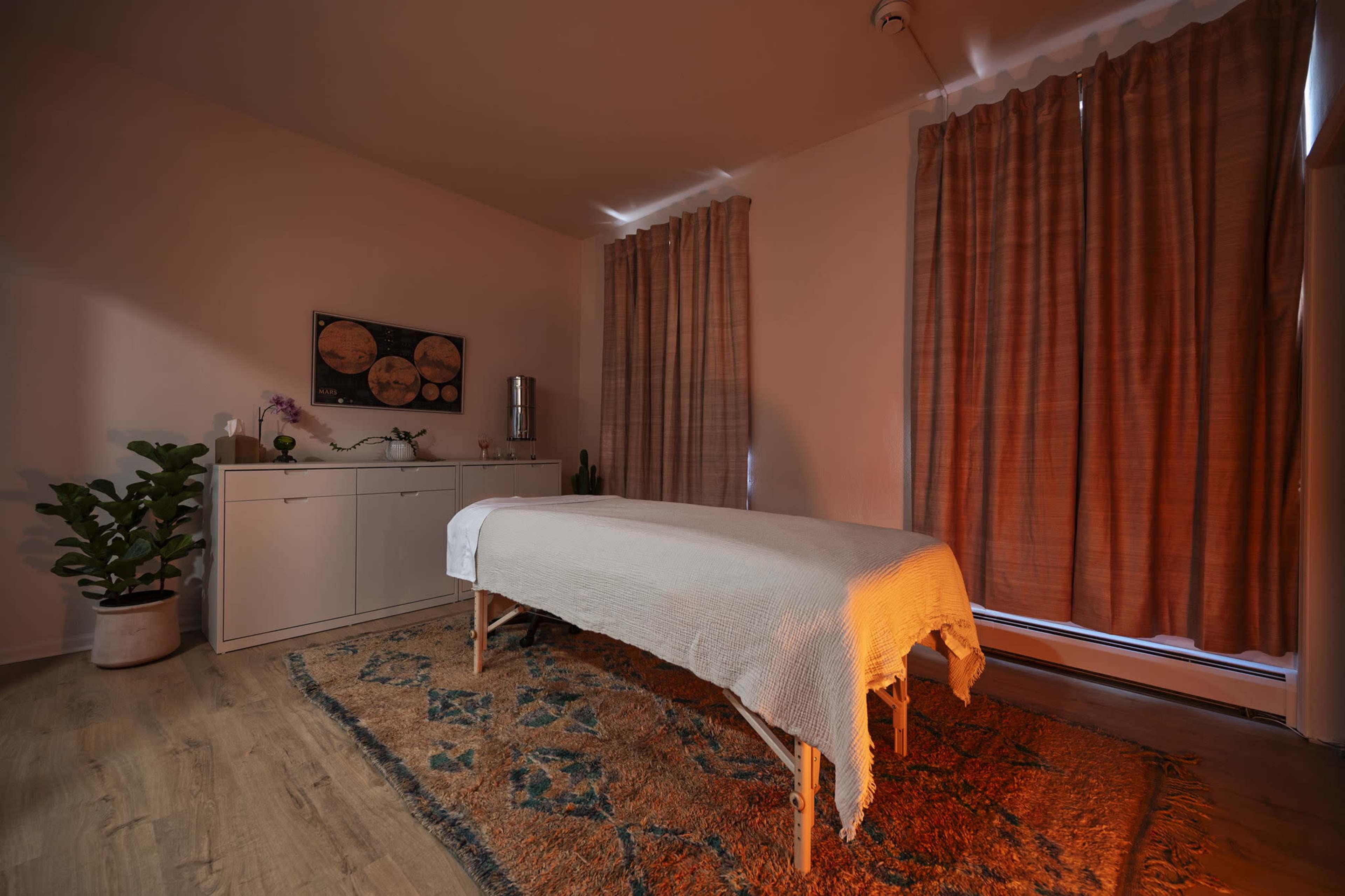 The image shows a softly lit room with a massage table covered by a white blanket, surrounded by curtains and a decorative rug.