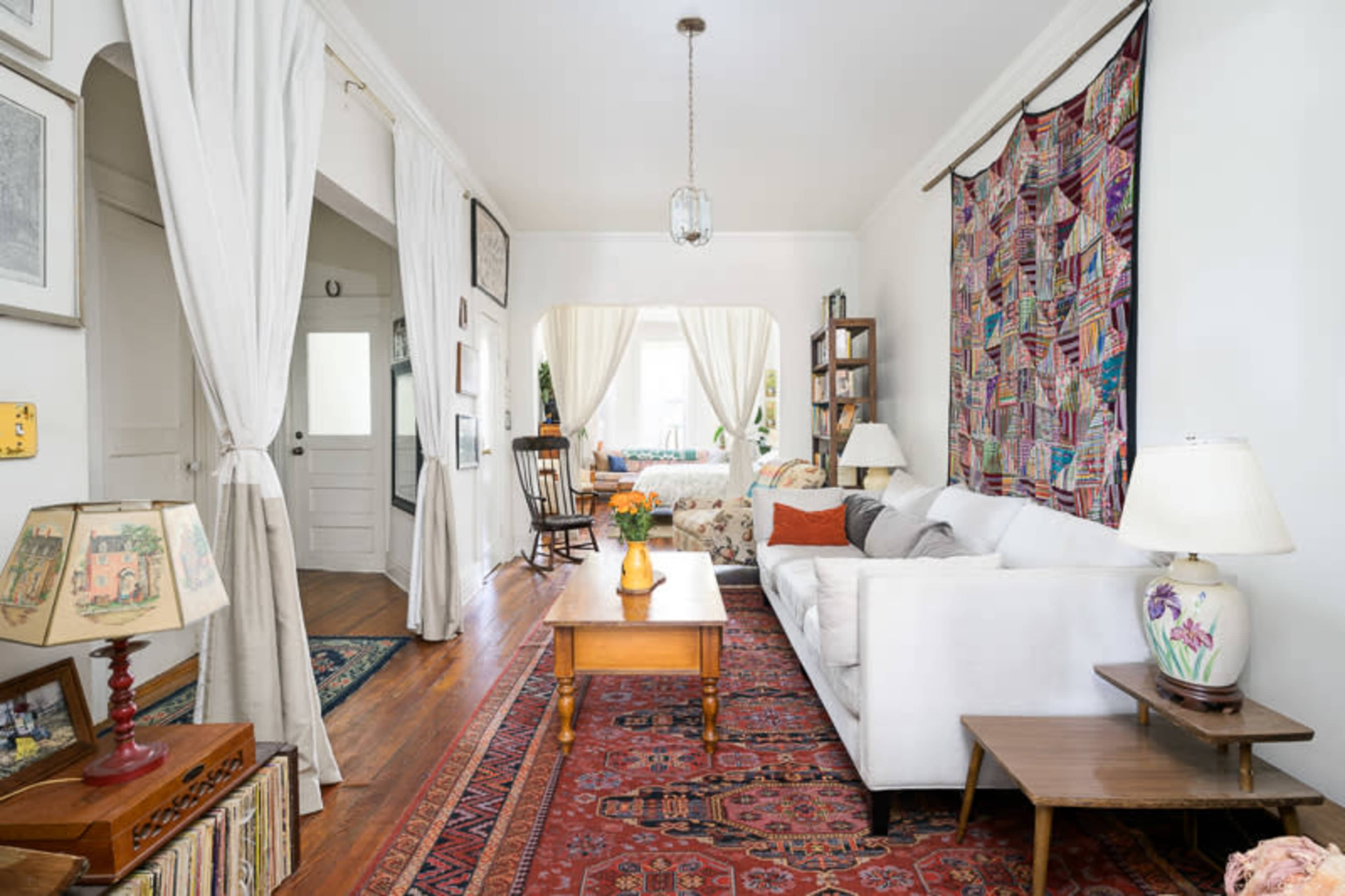 The image shows a cozy living room featuring a white sofa, a wooden coffee table, and decorative rugs, with light streaming in through large windows draped with curtains.