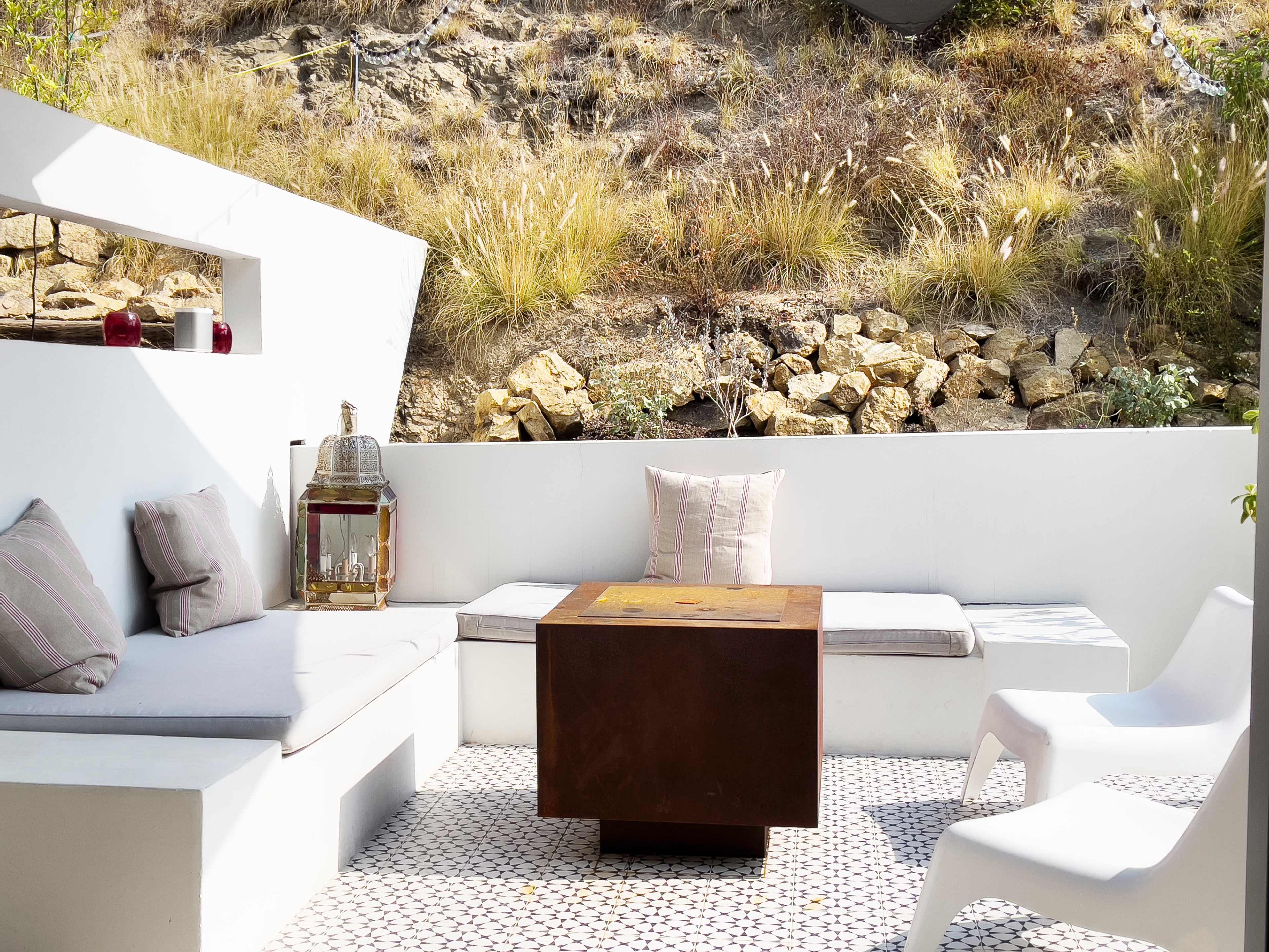 A modern outdoor seating area features a metal coffee table, surrounded by white couches and accented by decorative pillows, set against a backdrop of rocky terrain and dry grasses.