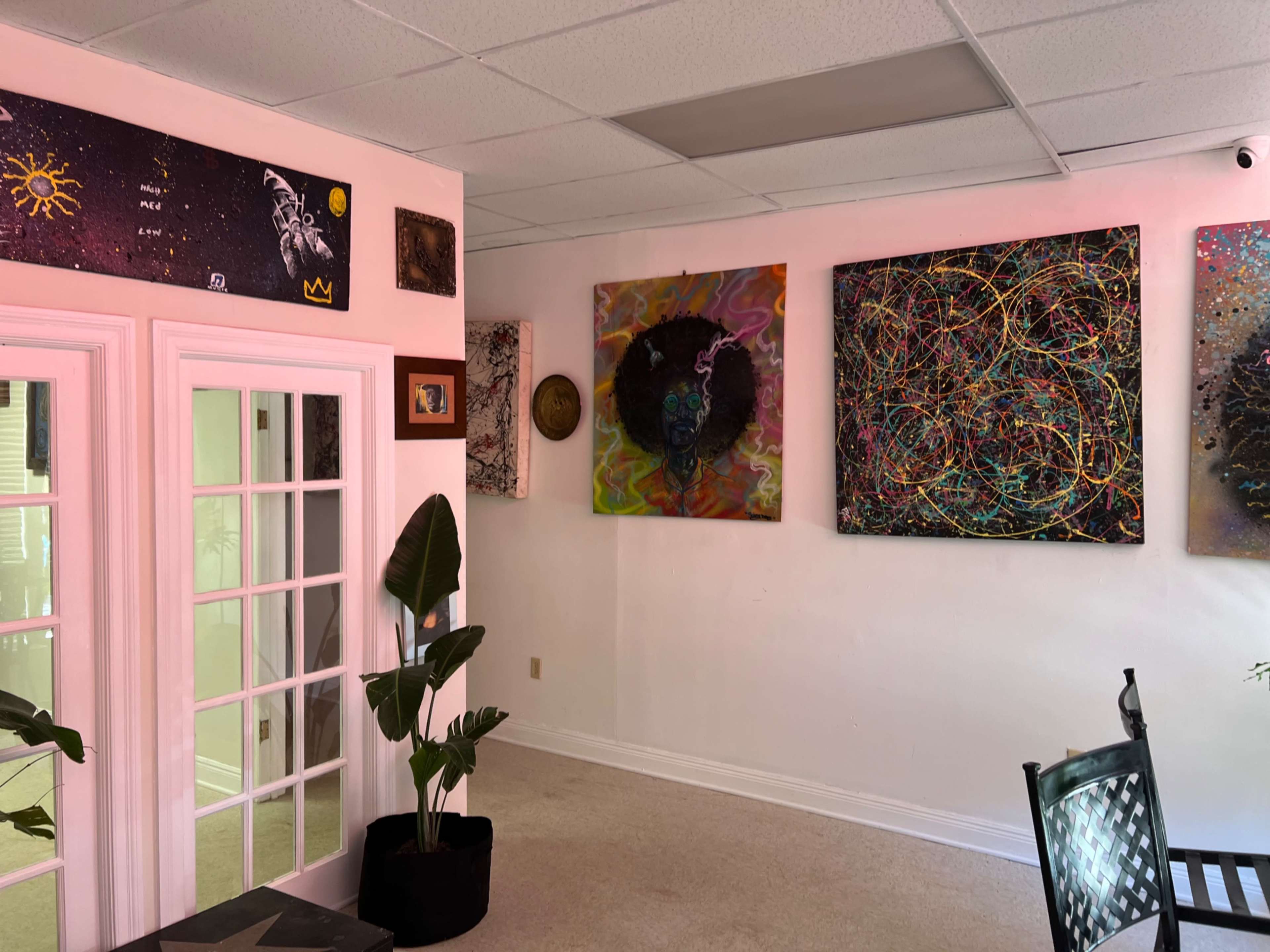 The image shows an interior space with colorful abstract artwork displayed on the walls, a potted plant in the foreground, and glass double doors leading to another area.