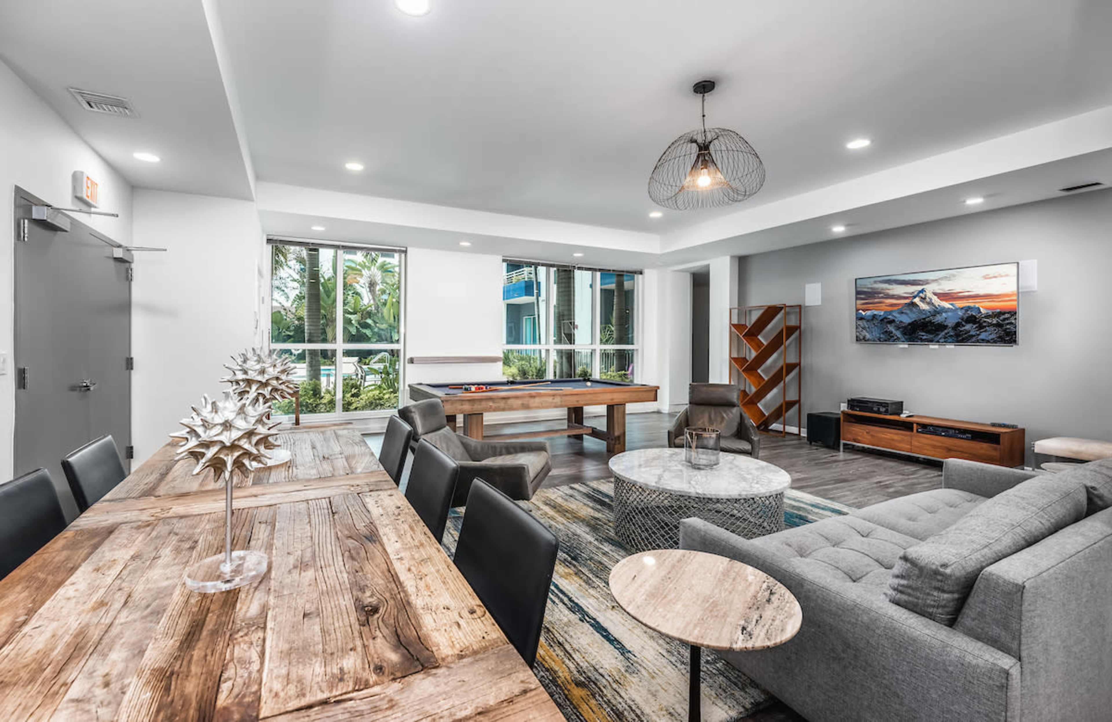 A modern lounge area featuring a pool table, a dining table with chairs, and a gray sofa, all illuminated by natural light from large windows.