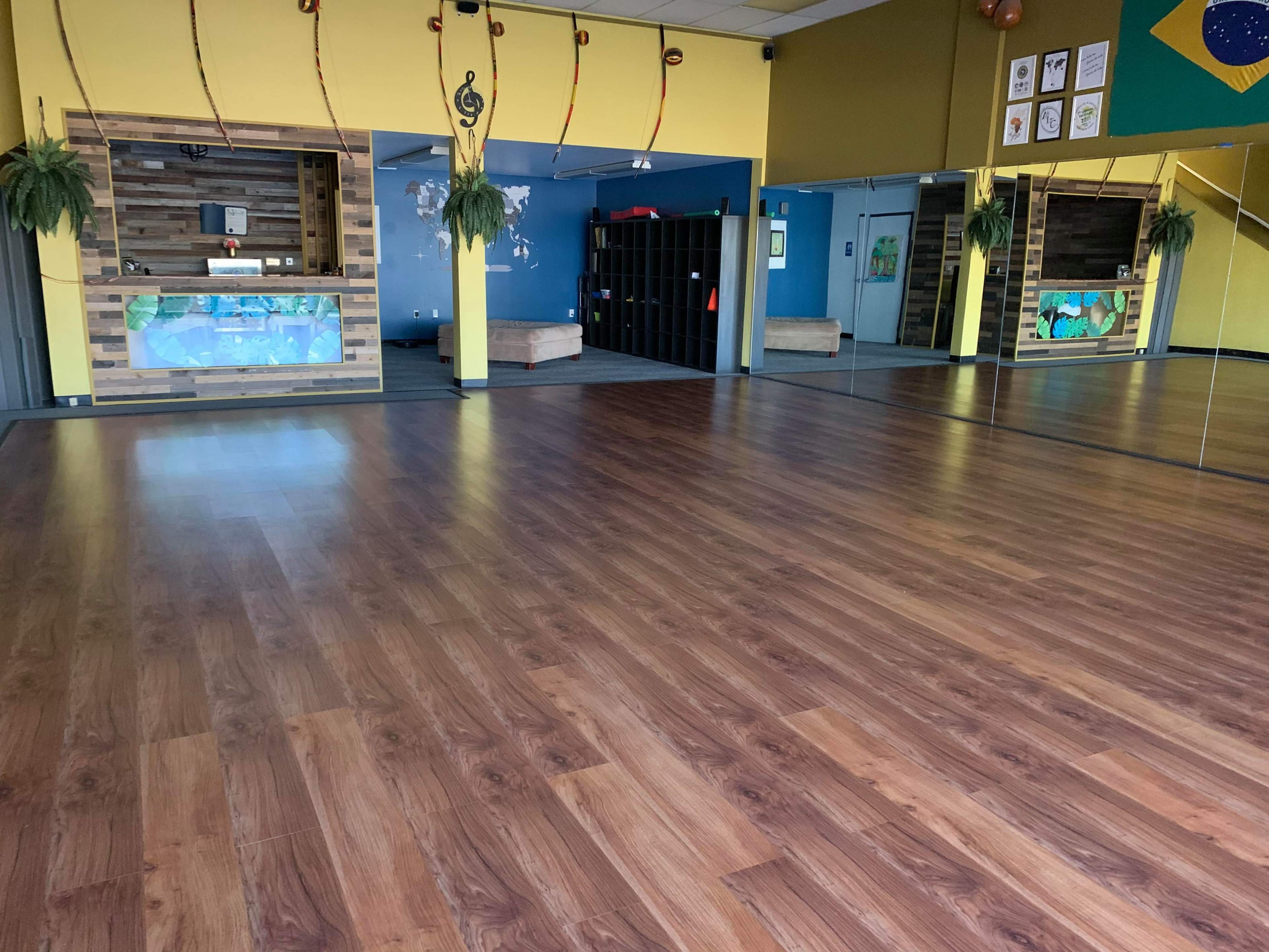 A spacious dance studio with wood flooring, mirrored walls, and a reception area featuring a colorful wall display.