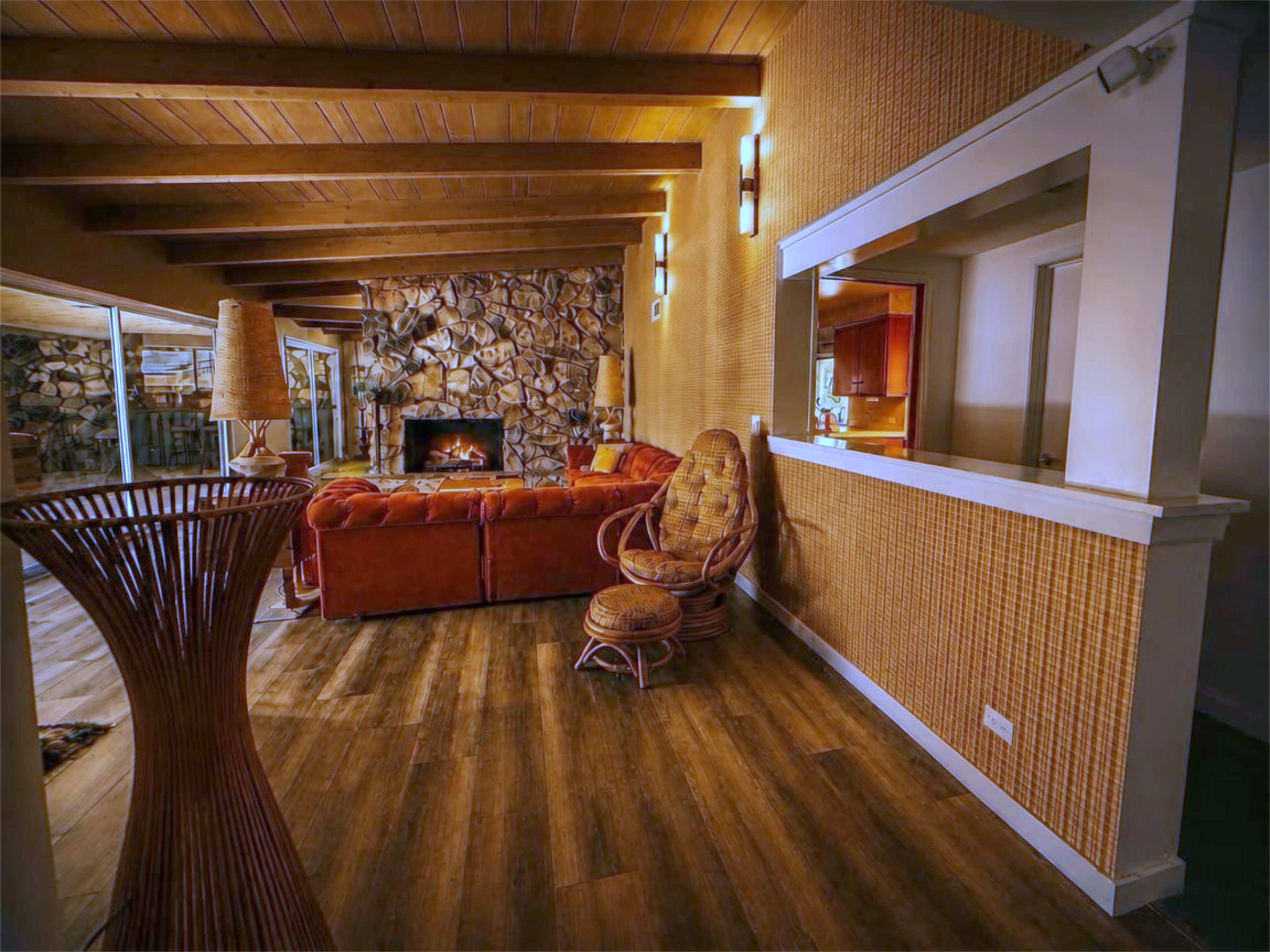 The image shows a living room with a stone fireplace, wooden beams on the ceiling, and a mix of wicker furniture and a large orange couch.