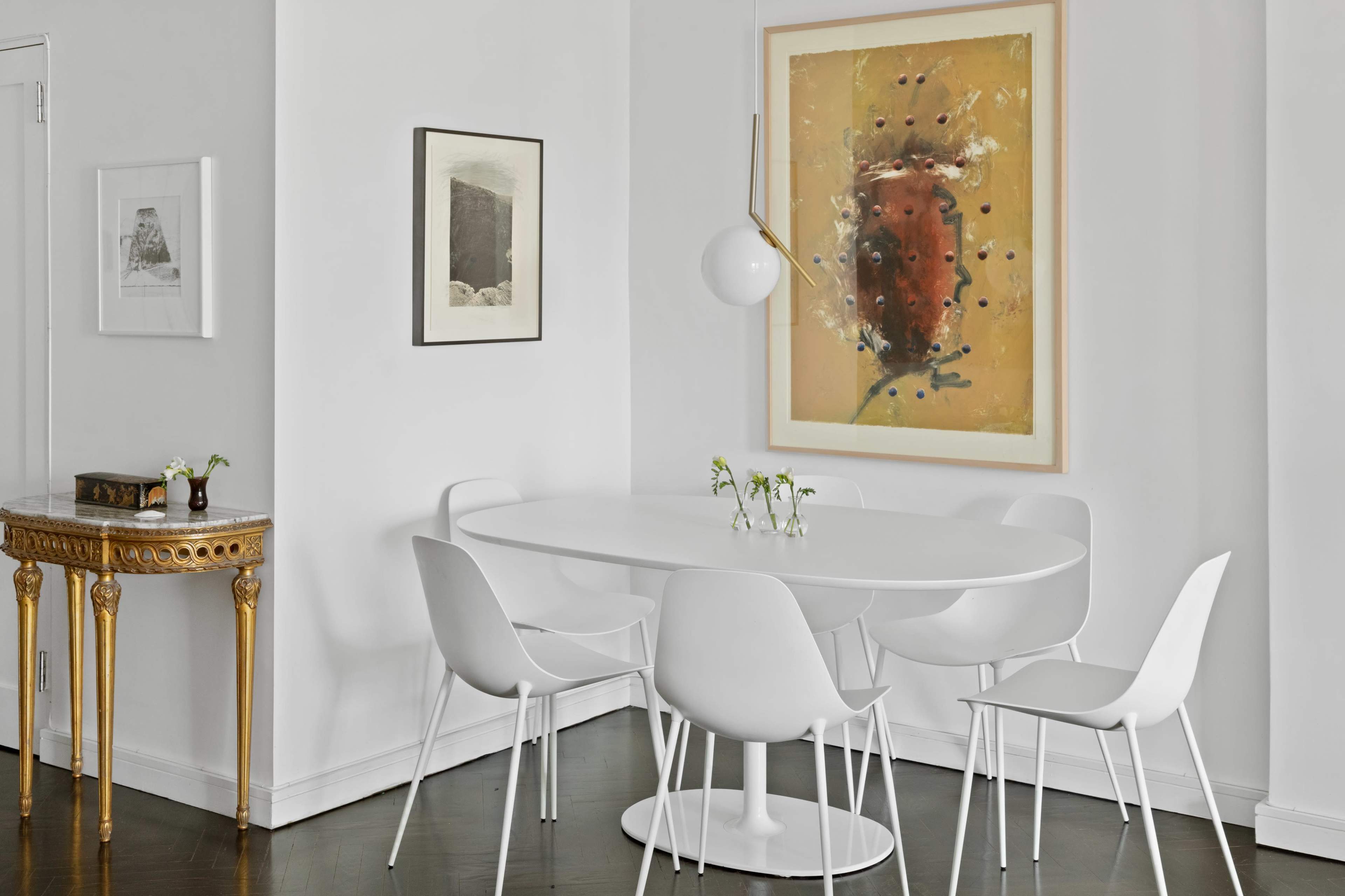 A round white dining table surrounded by white chairs is set against a wall featuring a large abstract painting and framed artwork.