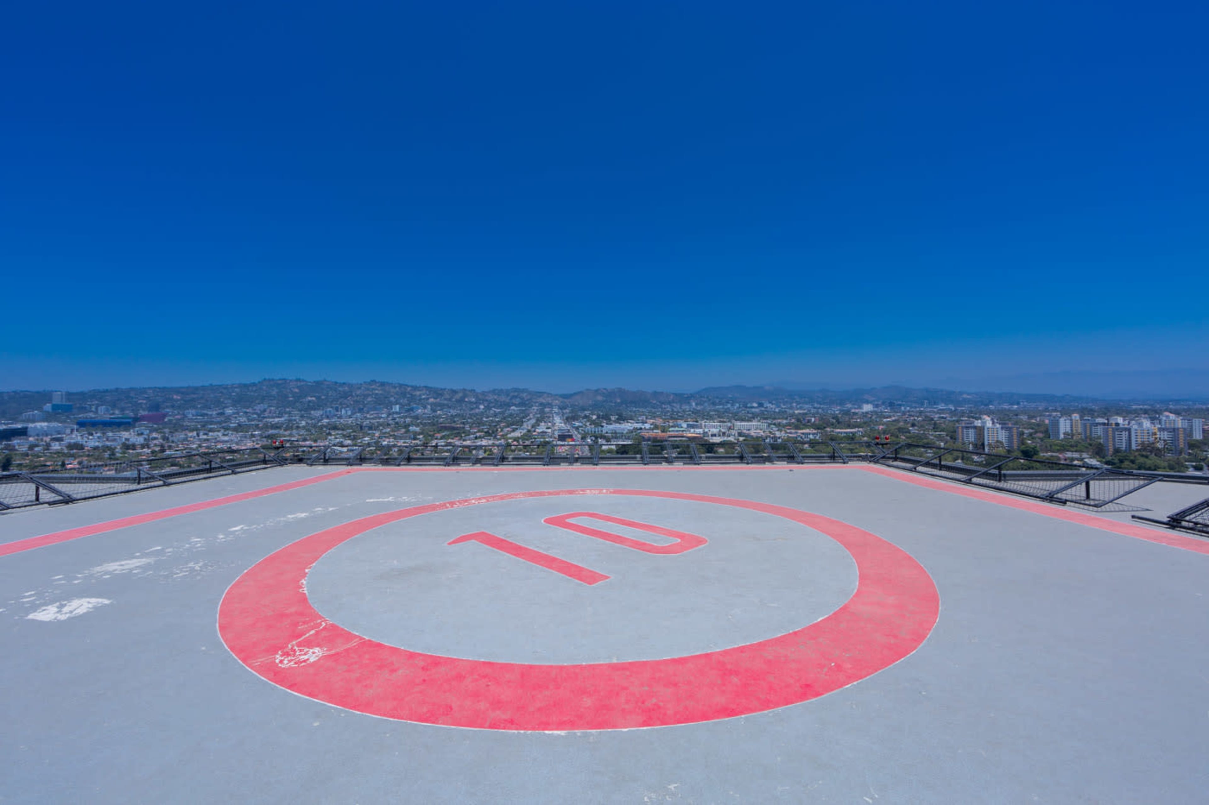 The image shows a rooftop helipad marked with a red circle and the number "10," overlooking a cityscape with distant hills under a clear blue sky.