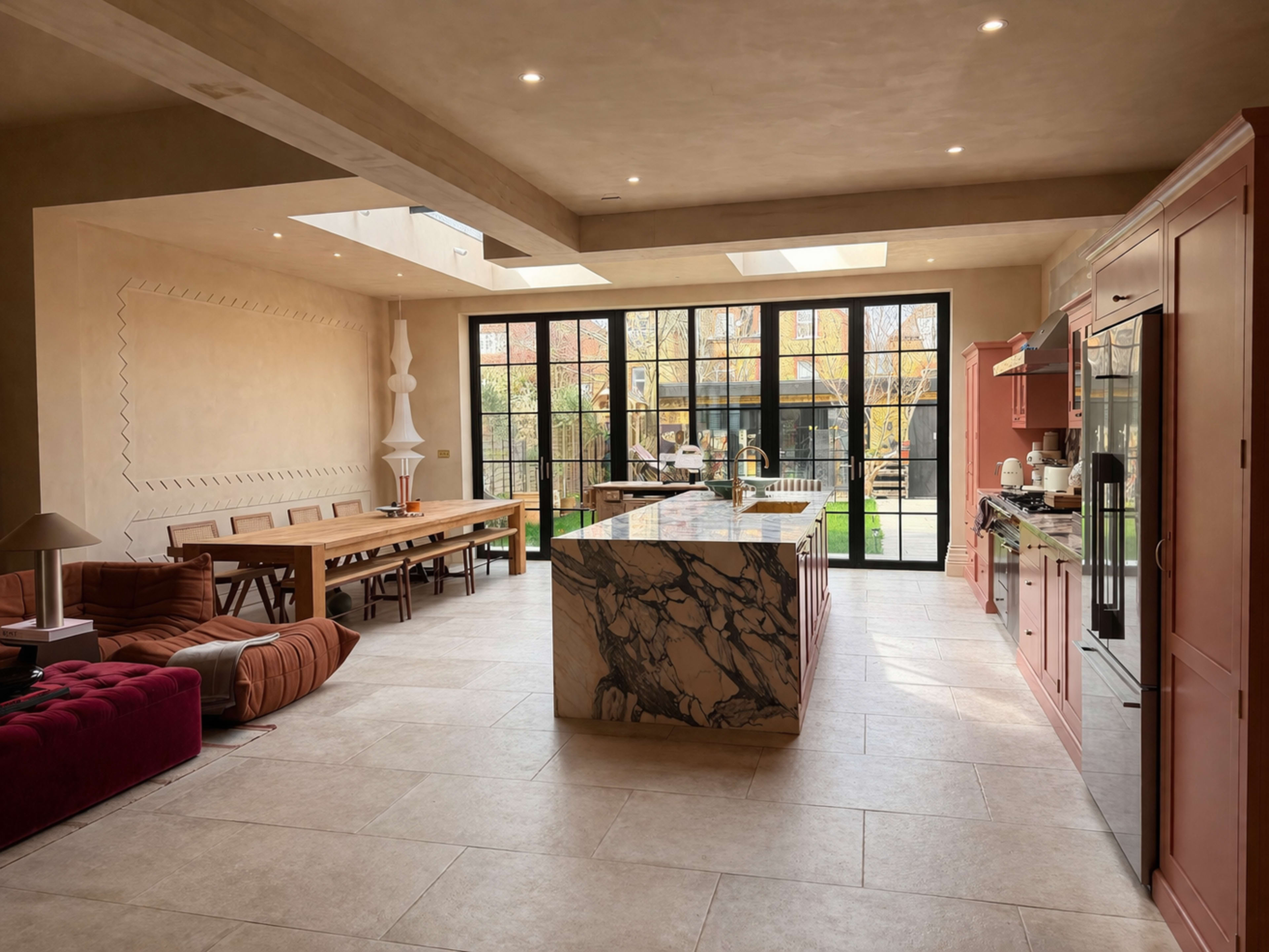 The image shows a spacious kitchen with a large marble island, open shelving, a dining area, and large glass doors that provide a view of a garden.