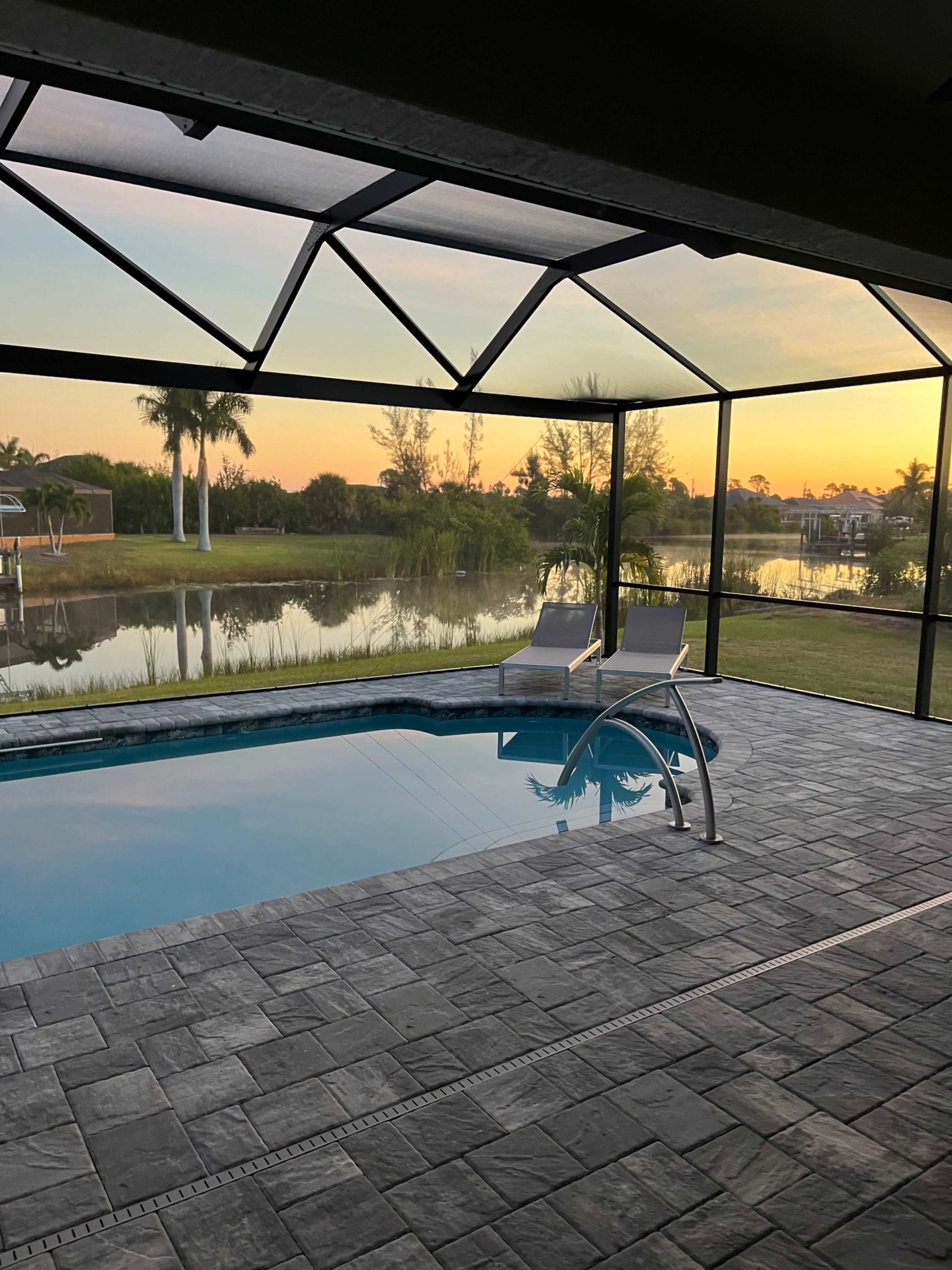 A swimming pool with two lounge chairs overlooks a calm lake at sunset, framed by a screened enclosure.