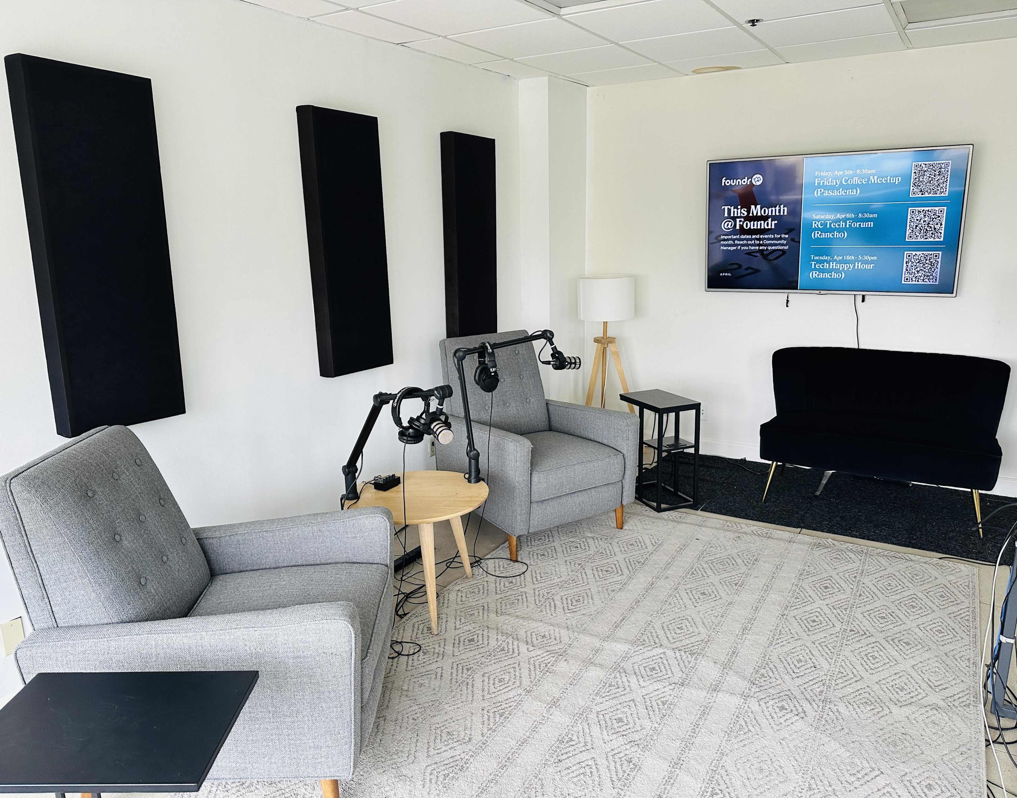 A recording studio with two gray chairs, a small wooden table, a black couch, and a wall-mounted screen displaying information, all set against a carpeted floor.