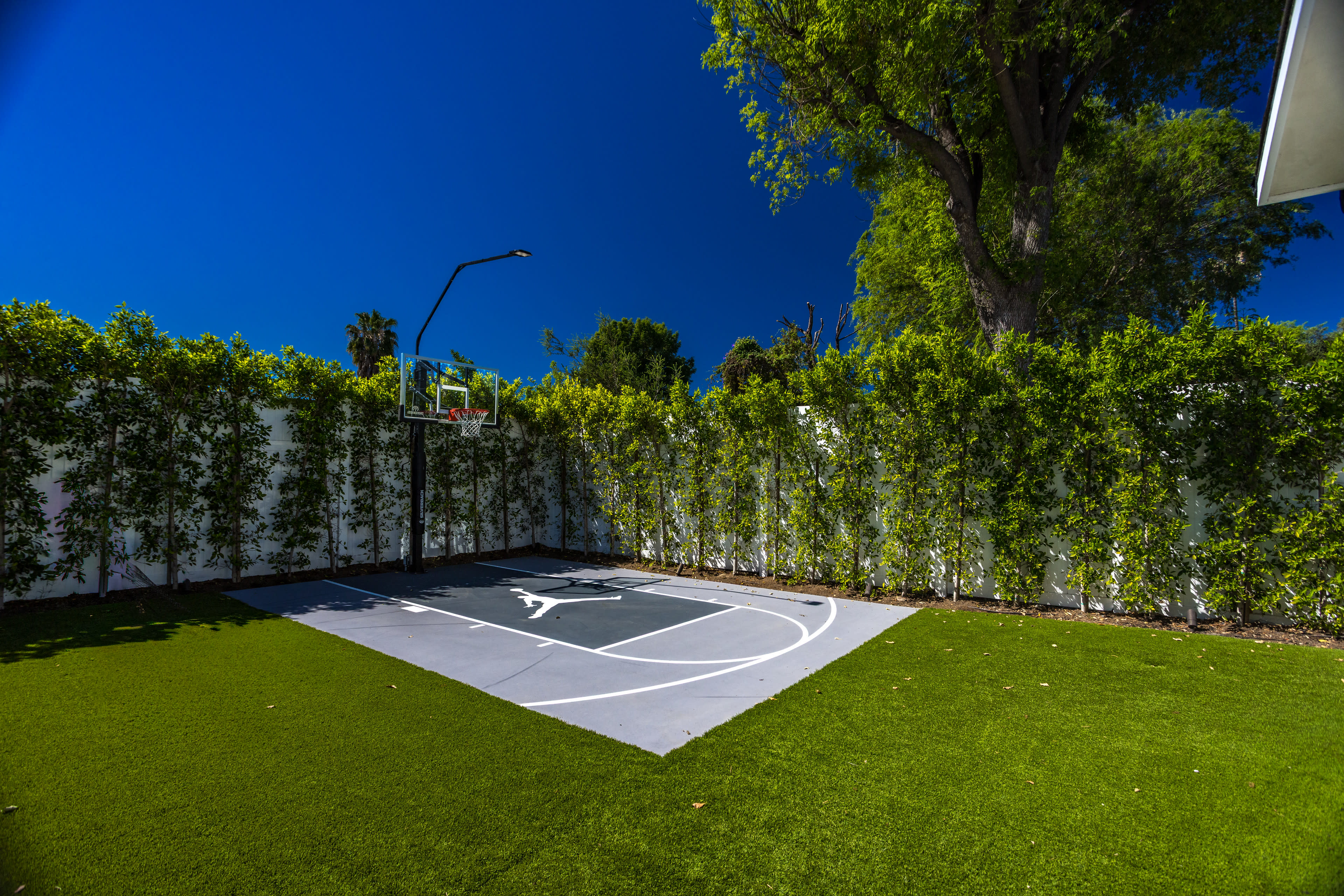 The image shows a small outdoor basketball court surrounded by tall green hedges and a clear blue sky.