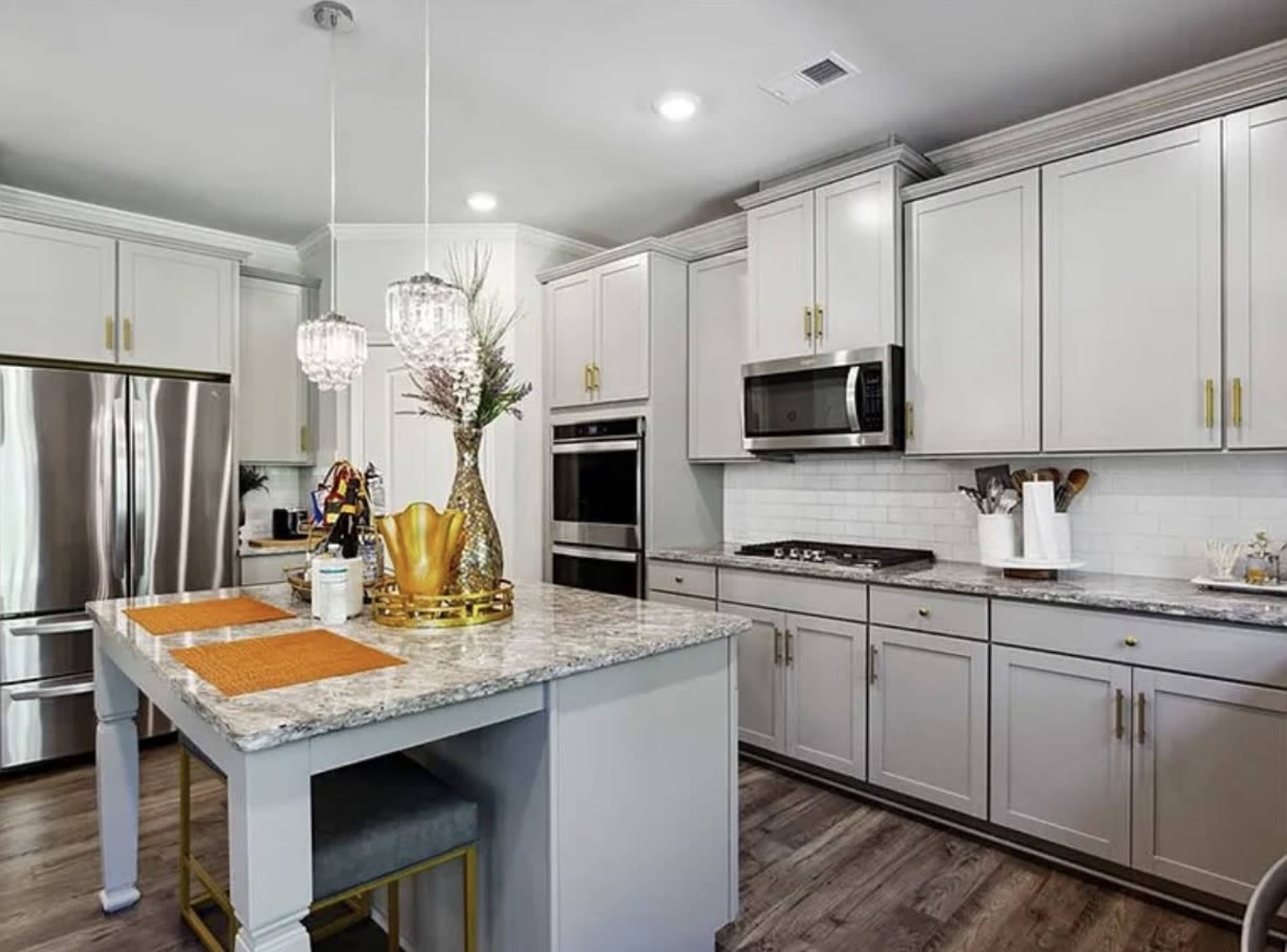 The image depicts a modern kitchen featuring gray cabinets, stainless steel appliances, a large island with a marble countertop, and decorative accents.