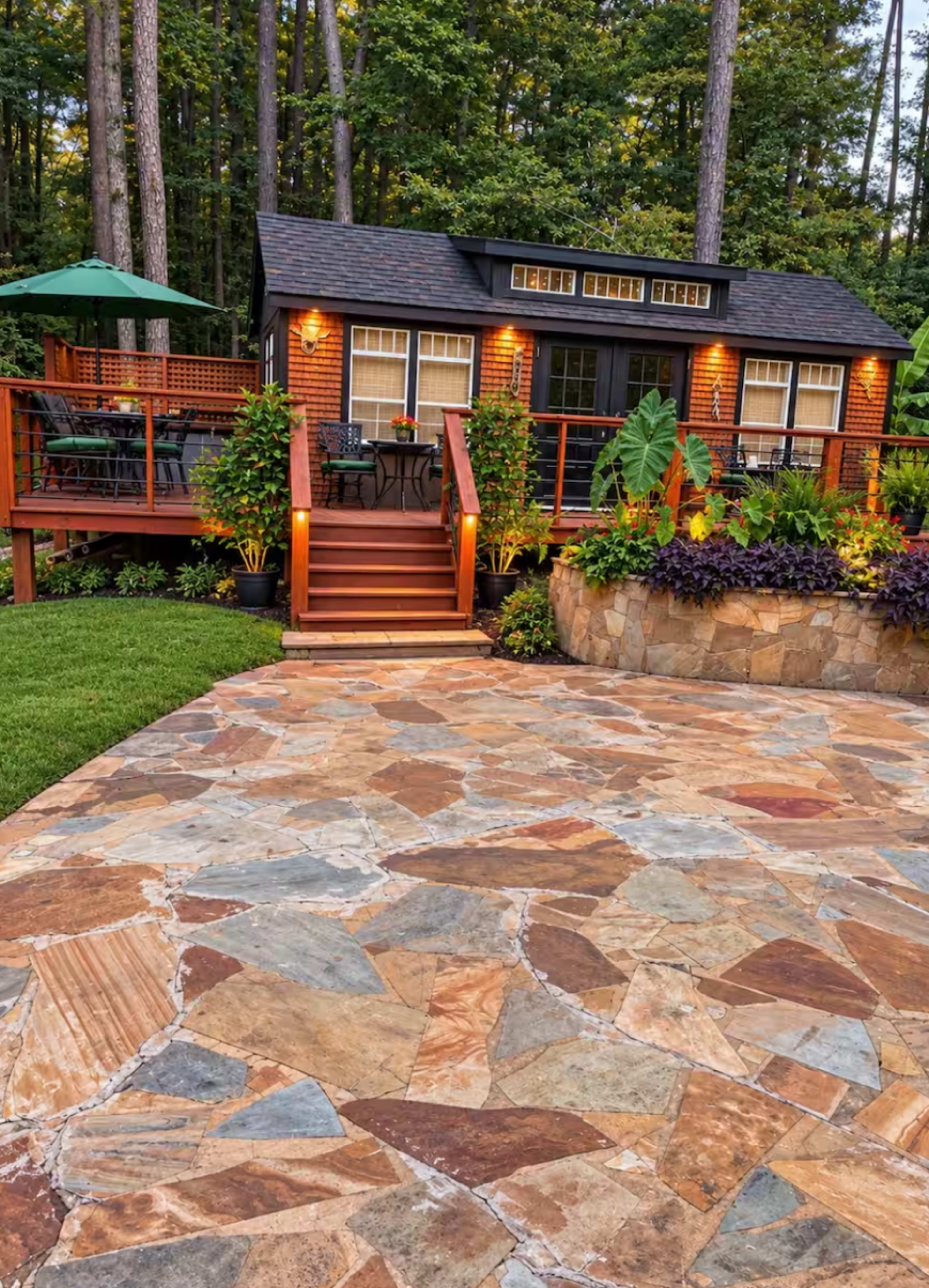 A wooden deck leads to a small house surrounded by greenery and a stone patio with various plants.