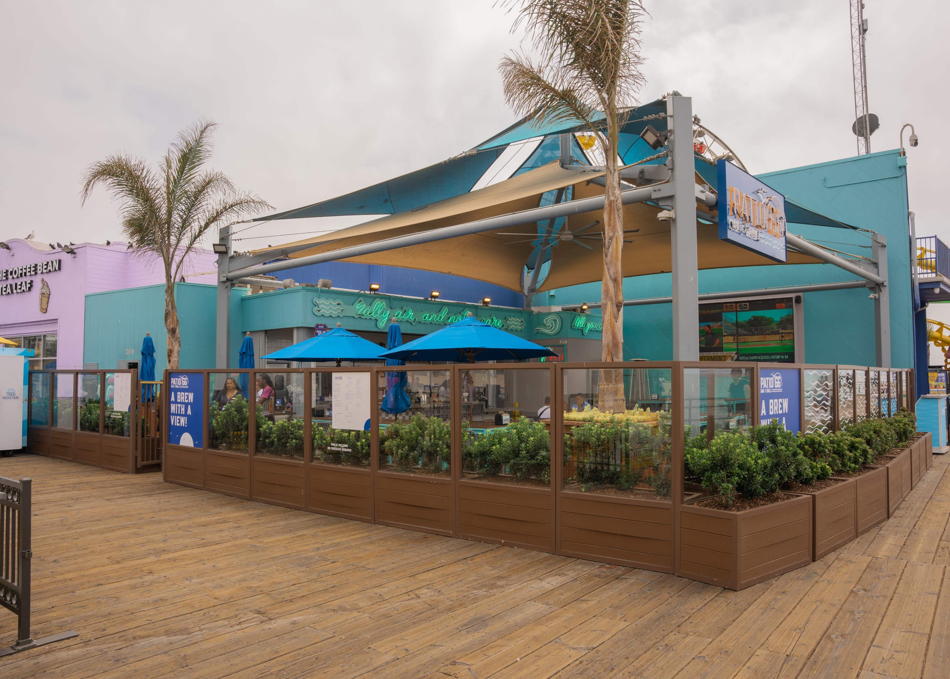 A restaurant with blue umbrellas is situated on a wooden boardwalk and features palm trees and a fenced outdoor seating area.