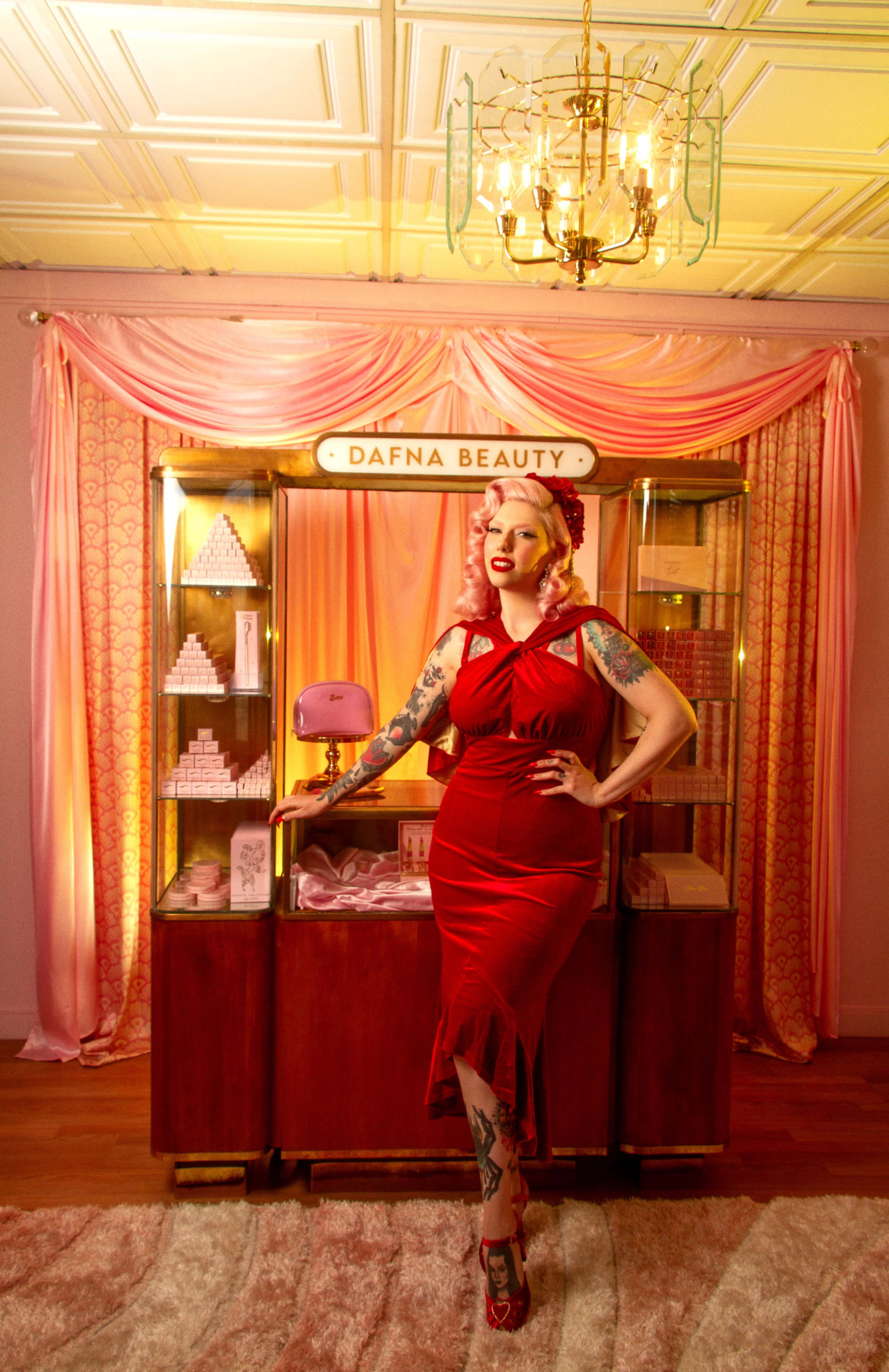 A woman in a red dress poses in front of a display of beauty products under a chandelier.
