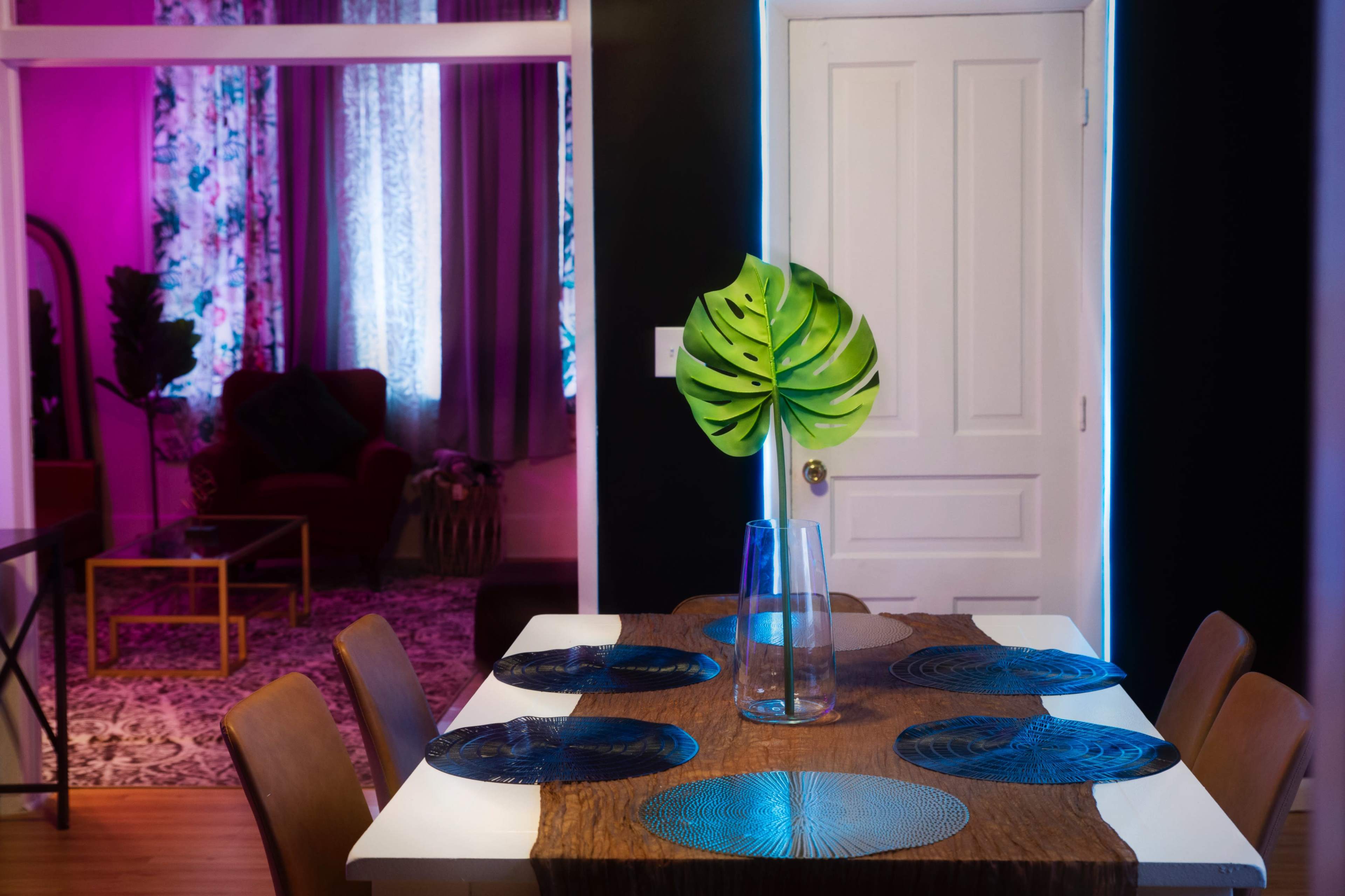 A dining table with blue plates and a vase containing a large green leaf sits in a room accented by purple lighting and floral curtains.