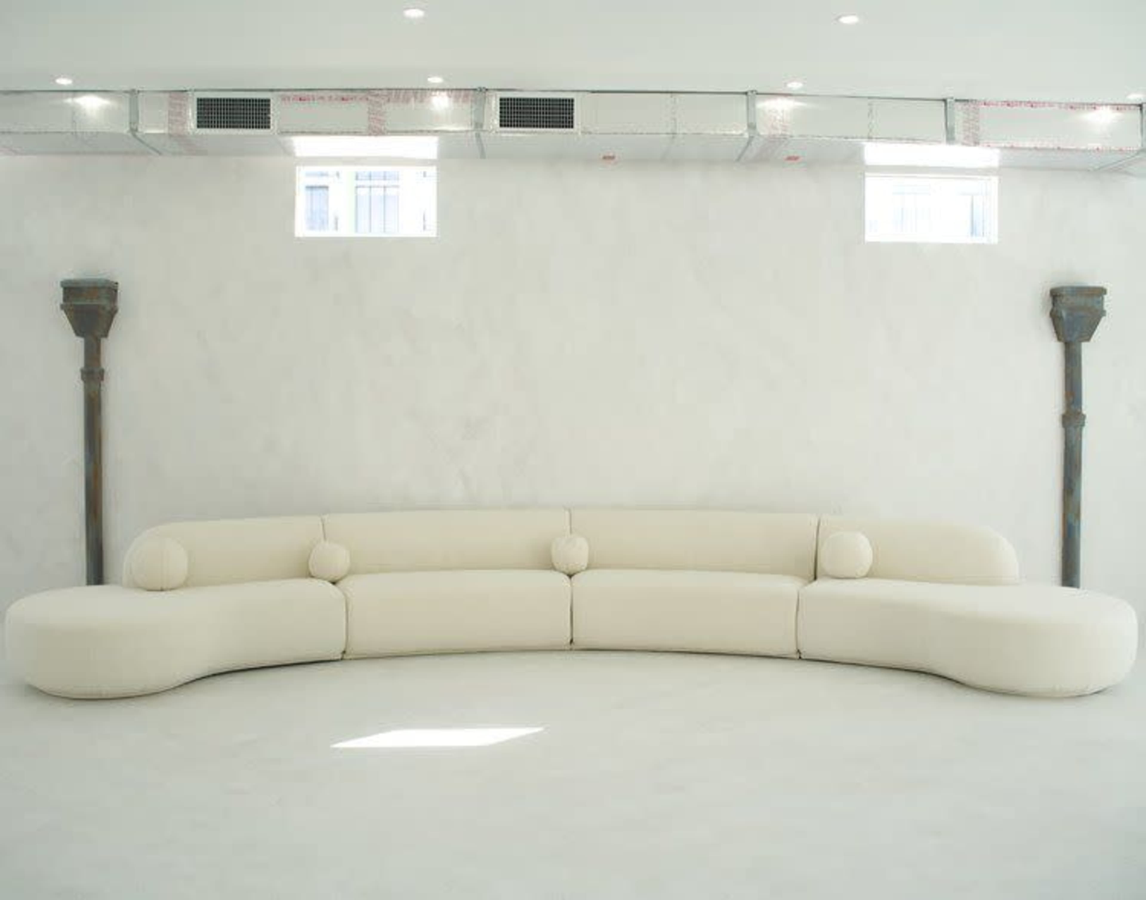 The image shows a curved, white, modern sofa positioned against a plain, light-colored wall with small windows and decorative supports.
