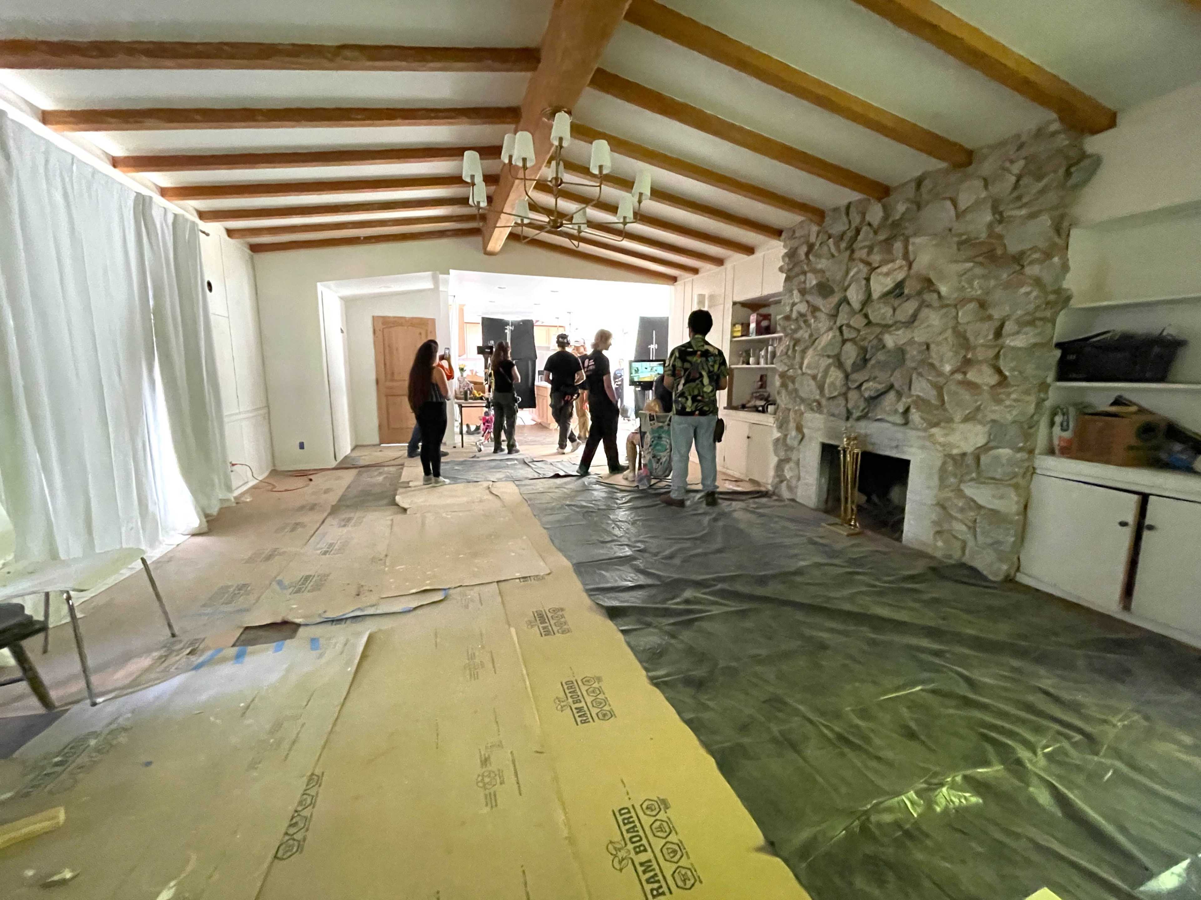 A group of people is working in a partially furnished room with exposed beams, a stone fireplace, and a covered floor.