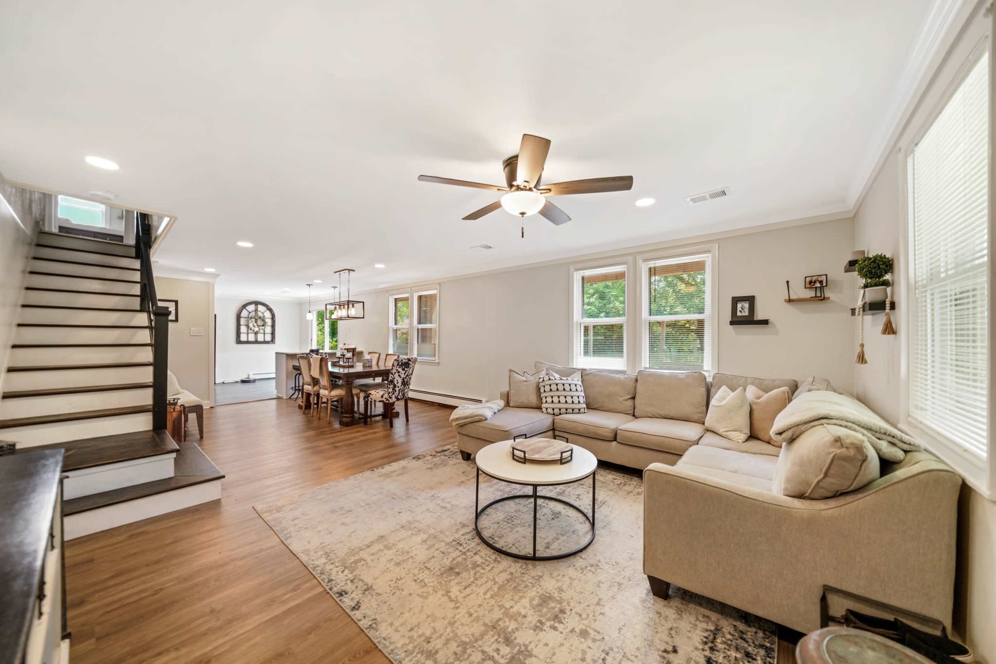 A spacious living room with a sectional sofa, a round coffee table, a ceiling fan, and a staircase leading to an upper level, with dining space visible in the background.