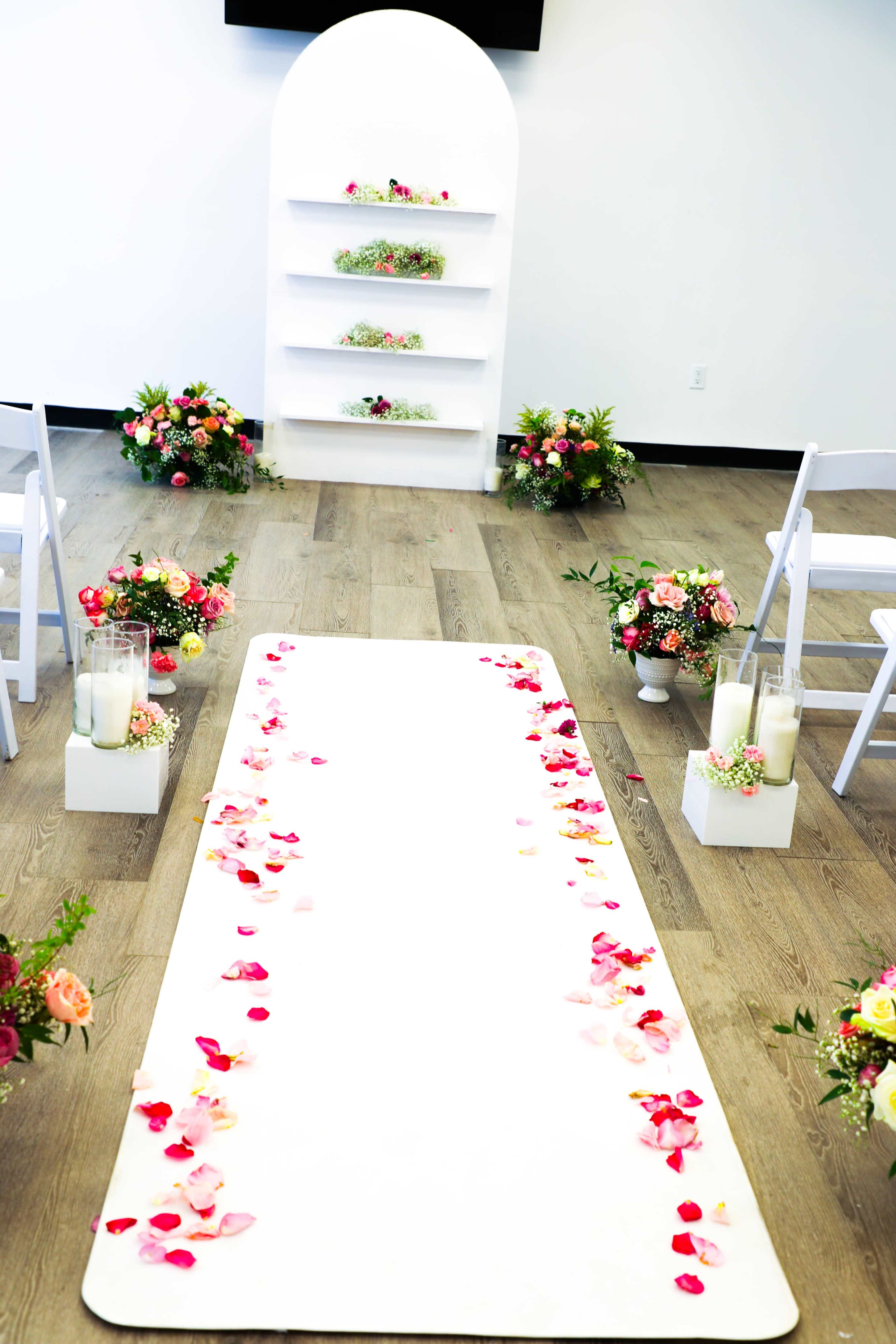 A decorated wedding ceremony setup with a floral aisle, white chairs, and a display shelf in the background.