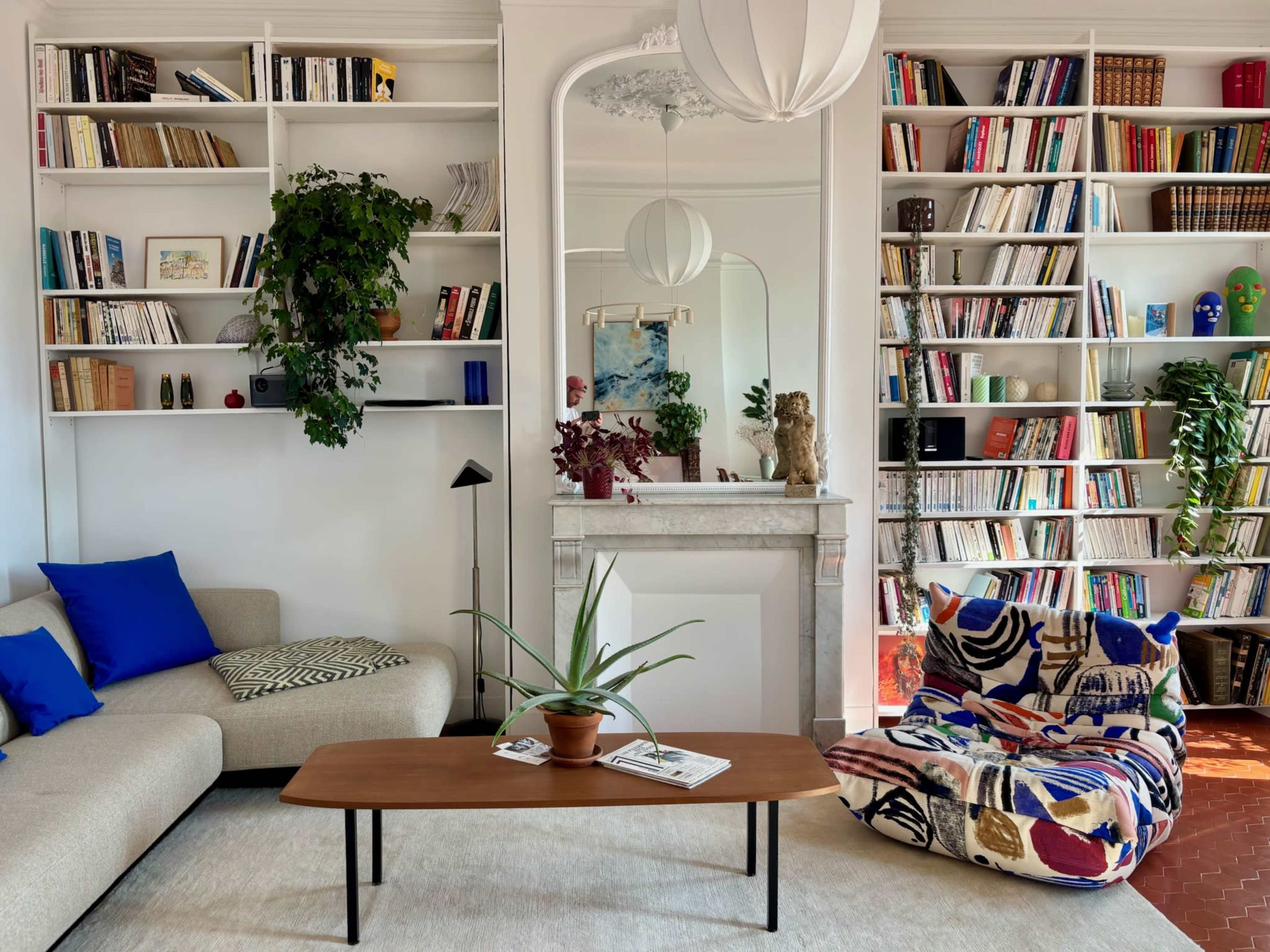 The image depicts a cozy living room with a beige sofa, a wooden coffee table, a bookshelf filled with books, and decorative plants.