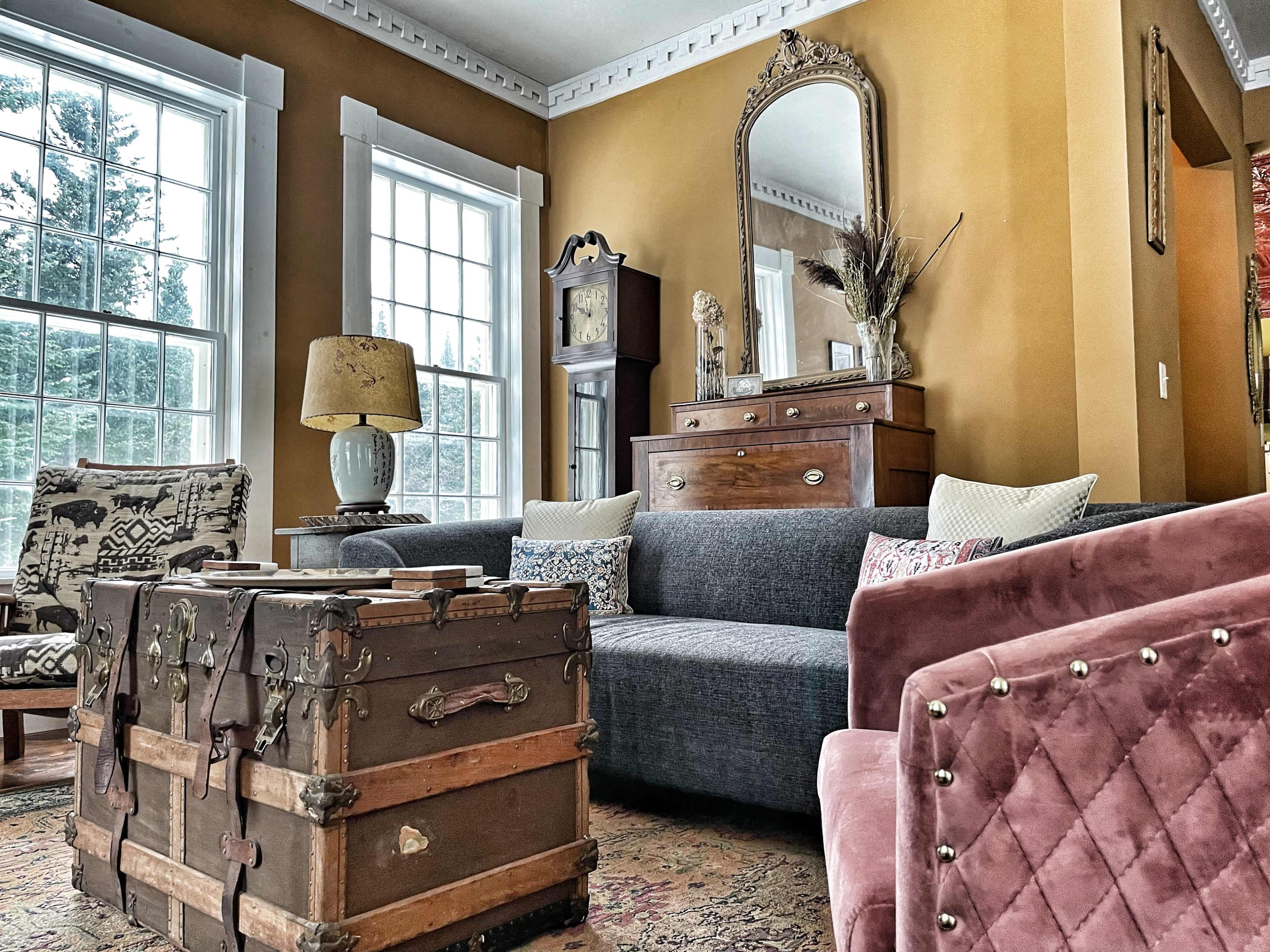 A living room features a gray sofa, vintage trunk, and a tall mirror framed in ornate gold, with large windows letting in natural light.