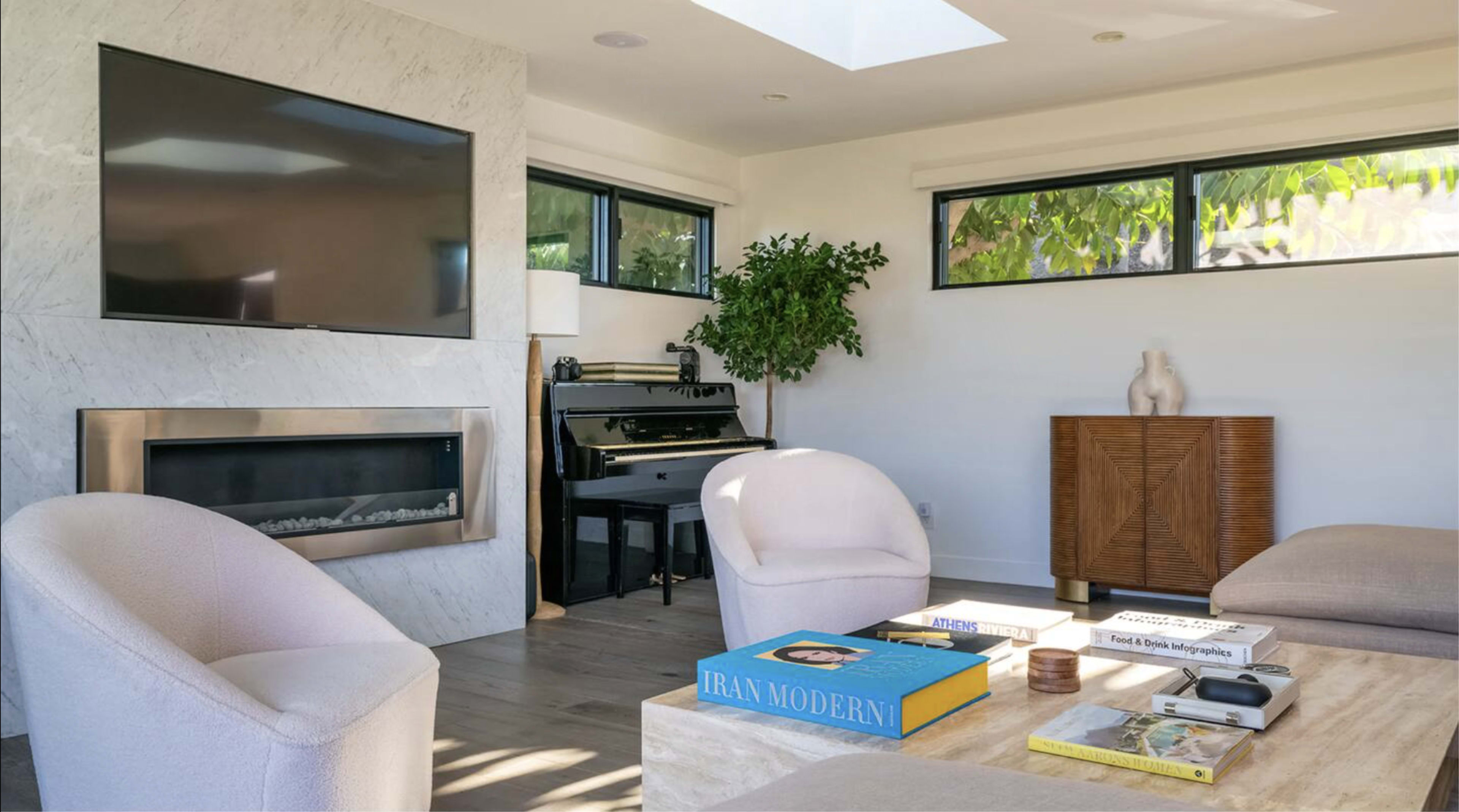 A modern living room features a television mounted on a marble wall, a piano beside large windows, and a coffee table with books and decorative items.