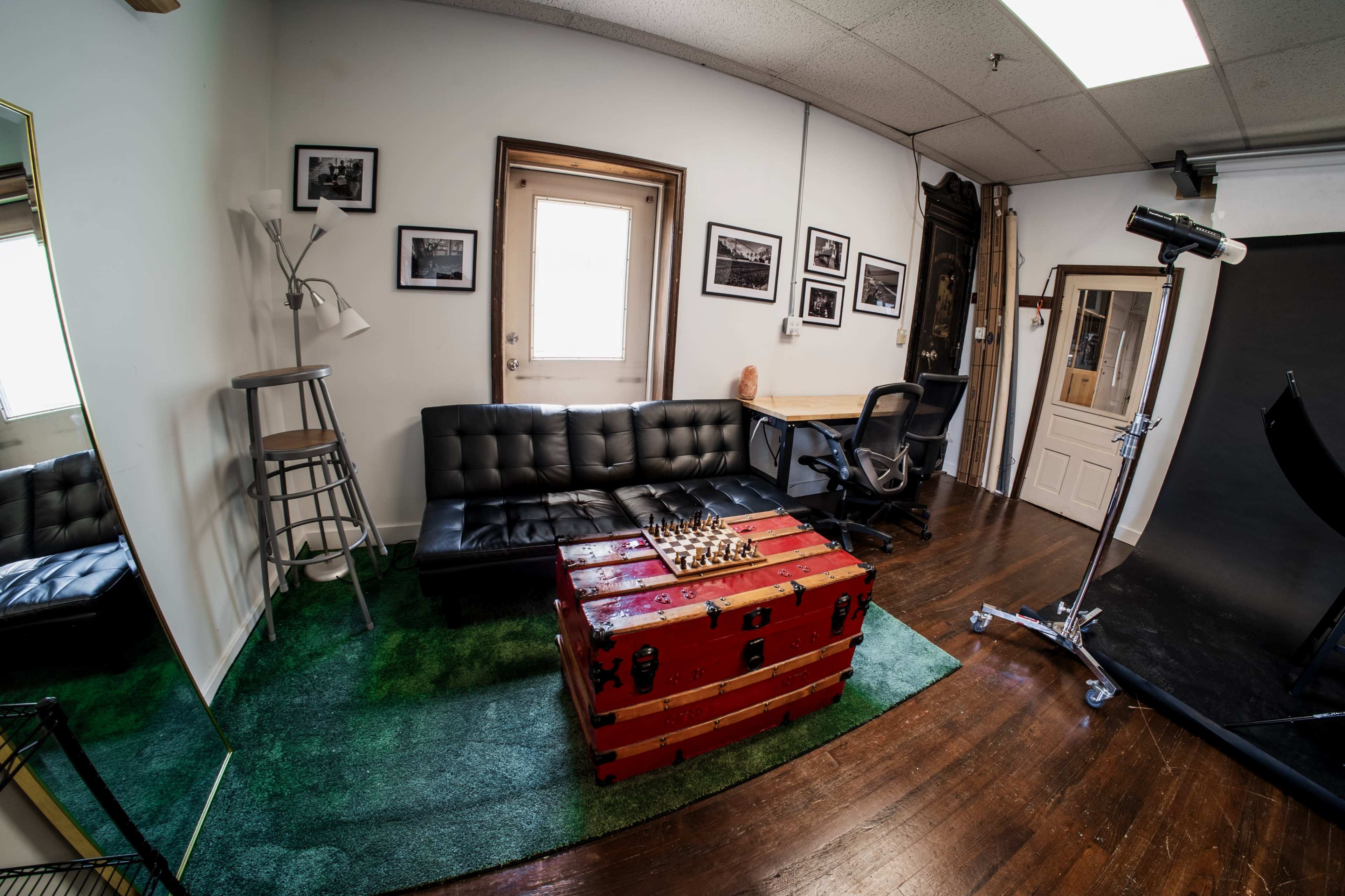 The image shows a room with a black sofa, a wooden chest, a chair, and various framed photographs on the walls.