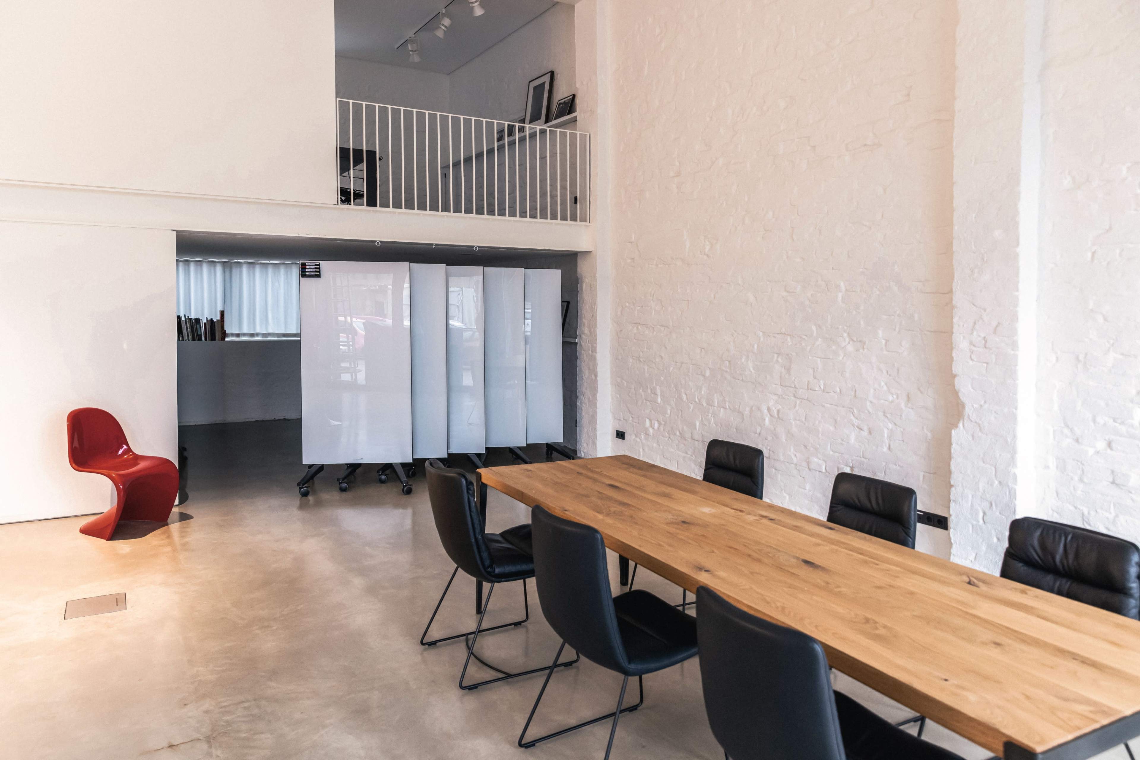 The image shows a modern office space featuring a wooden conference table surrounded by black chairs, with a red chair in the corner and a white partitioned area in the background.