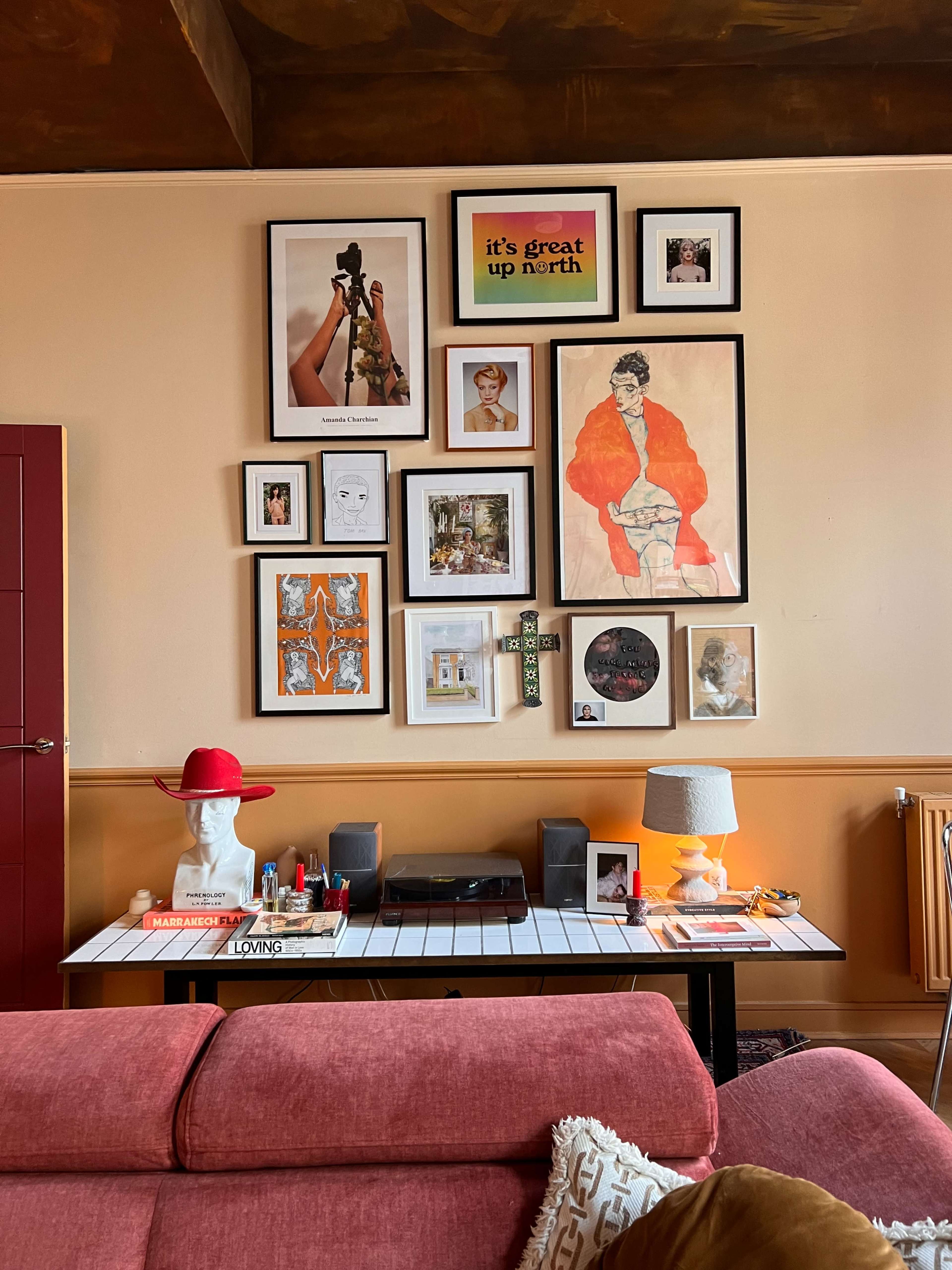 The scene features a living room wall adorned with various framed artworks and photographs, complemented by a white table displaying decorative items and a record player.