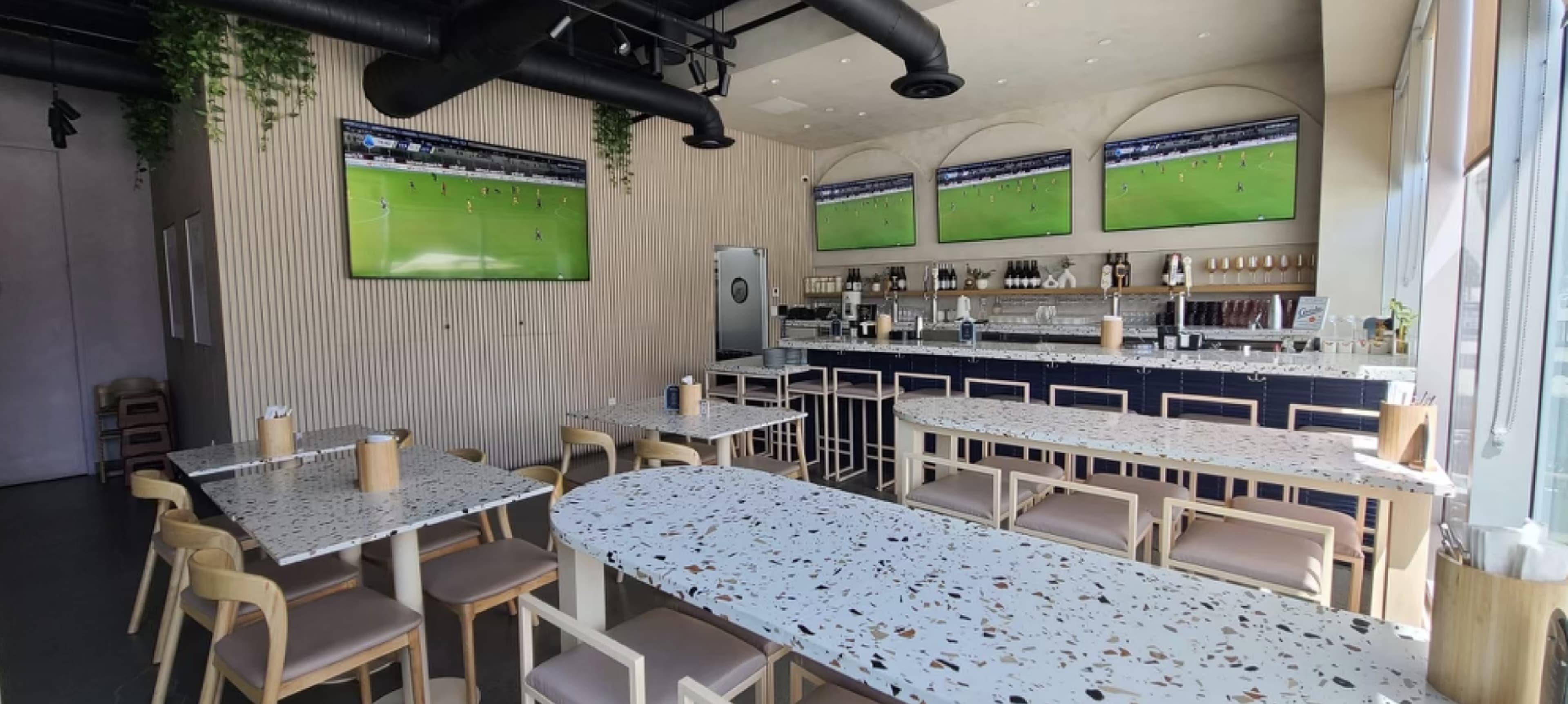 The image shows a modern restaurant interior featuring several tables, a bar area with drinks, and multiple screens displaying a sports event.