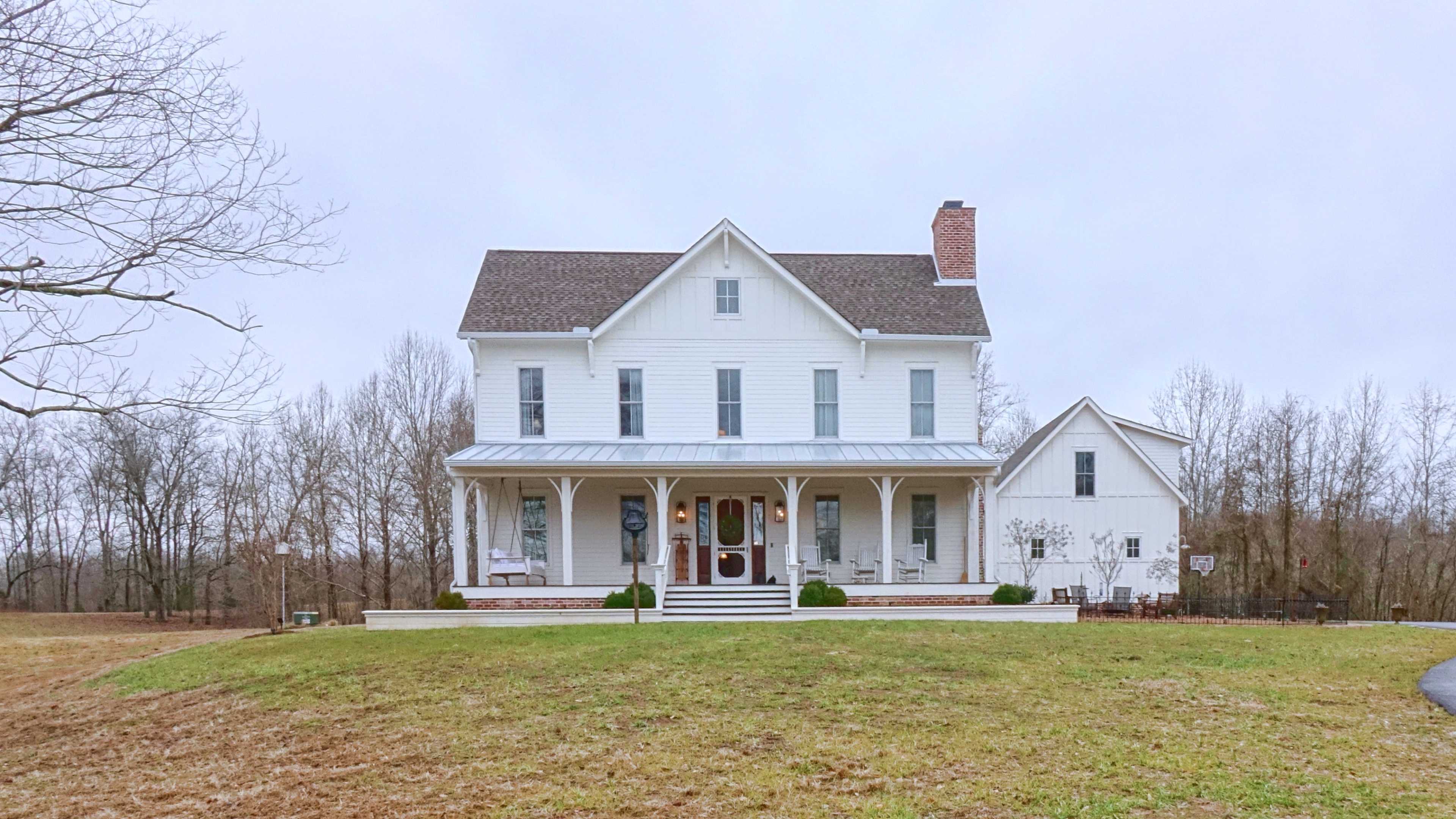 A large, two-story white farmhouse with a covered porch and a symmetrical layout, surrounded by a grassy area and trees in the background.