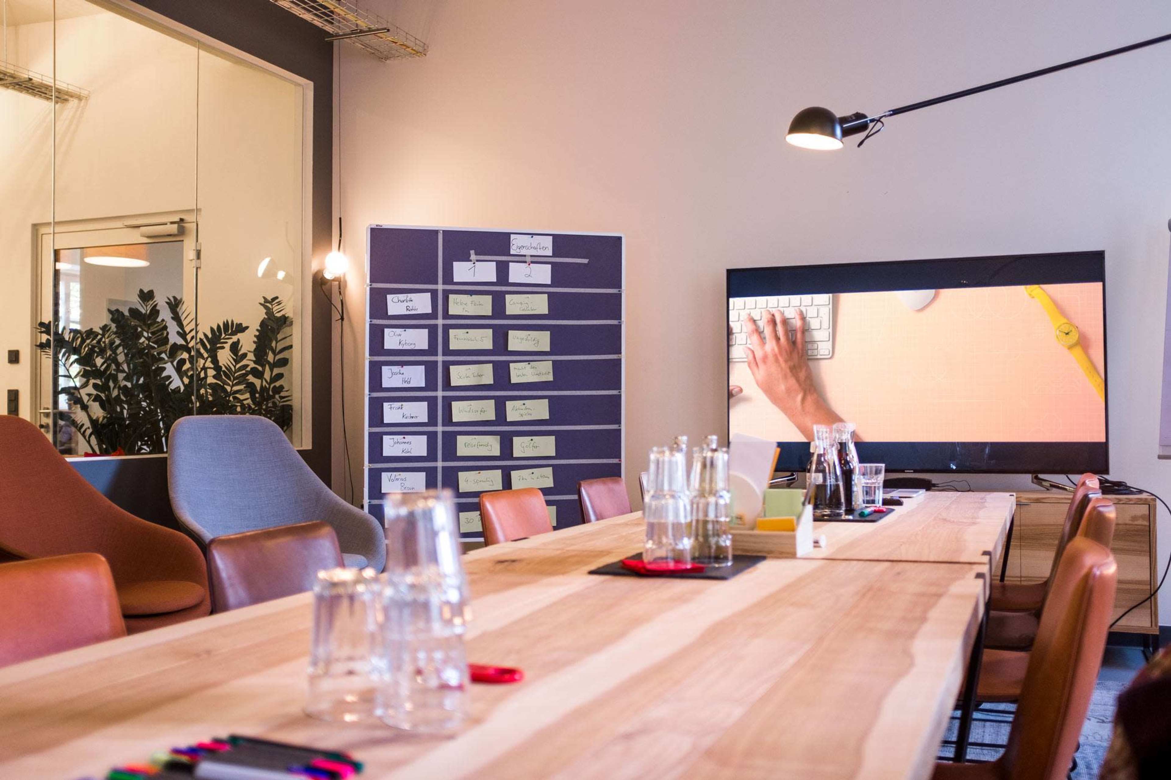 A modern conference room with a long wooden table, comfortable chairs, a large screen displaying a keyboard, and a wall with organized notes and charts.
