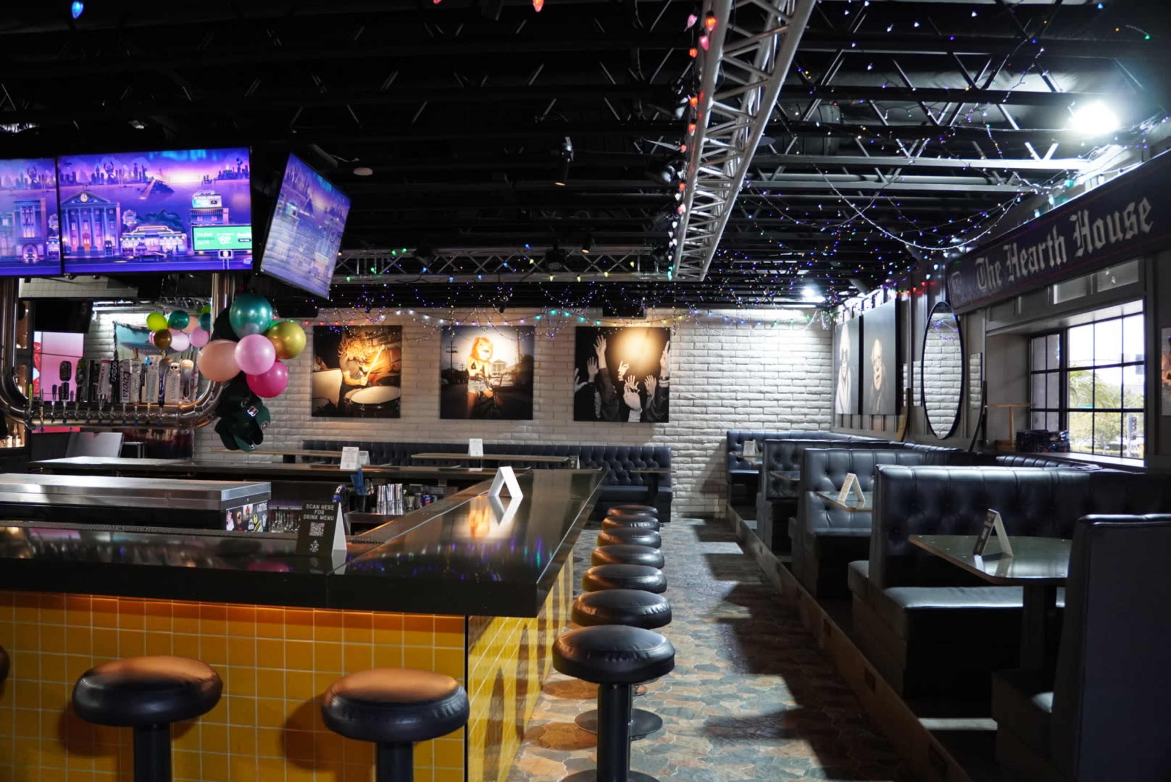 The interior of a restaurant features a bar with stools, black booths, and framed photographs on the walls, illuminated by string lights.